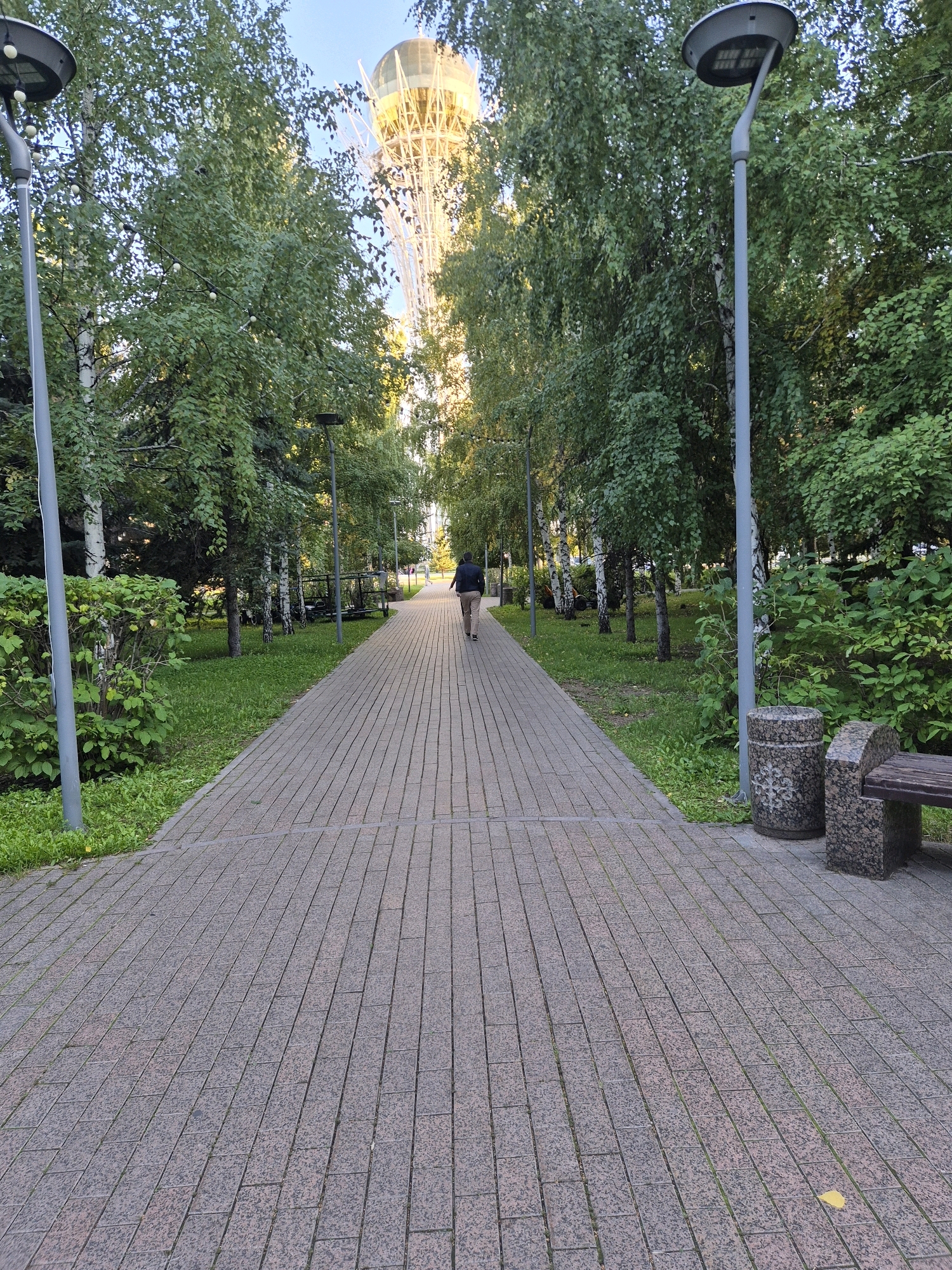 The image captures a paved walkway lined with trees, leading towards a distinctive tower with a spherical top. The walkway is constructed from rectangular stone tiles with a reddish-brown hue. On either side of the path, lush green grass and various shrubs and trees are visible. Modern streetlights with circular lamps are positioned along the walkway. In the foreground, to the right, a stone bench and a decorative stone trash receptacle are present. A single person, dressed in a dark jacket and light-colored pants, is walking away from the camera down the path. The background reveals the upper portion of a tall, white lattice tower topped with a golden, translucent sphere, identifiable as the Bayterek Tower in Astana, Kazakhstan. The sky is a clear, pale blue.
