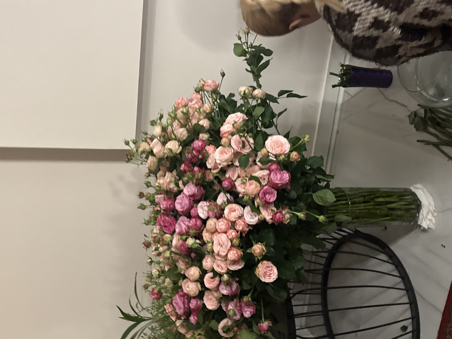 A large bouquet of pink and purple miniature roses dominates the scene, filling a black wire basket. Behind the flowers, a clear glass vase containing only stems lies horizontally. A person with blonde hair is partially visible at the top. In the background, a white wall and a glimpse of a patterned garment add to the composition. Additionally, other items like a purple vase and clear glass vase are on the table.