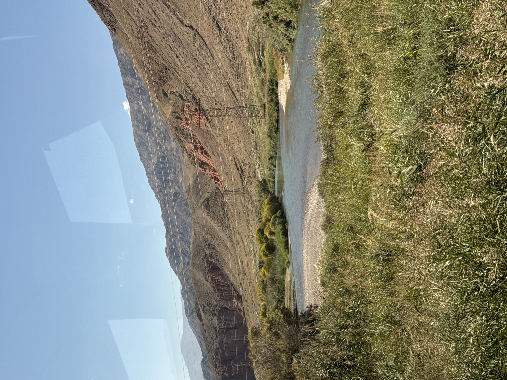 An aerial view captures a wide, shallow river flowing through a dry, mountainous landscape under a clear blue sky. The river's clear, turquoise water contrasts with the pale, pebble-strewn riverbed. Lush green reeds and tall grasses line the foreground, creating a dense natural barrier. On the far bank, a row of bright green, bushy trees stands out against the arid terrain. Beyond the trees, the rugged mountains rise steeply, their slopes covered in dry, brown vegetation and accented by striking red rock formations. Utility poles and power lines are visible, suggesting human presence in this otherwise wild environment. The scene is partially obscured by reflections on the surface, possibly from a window.