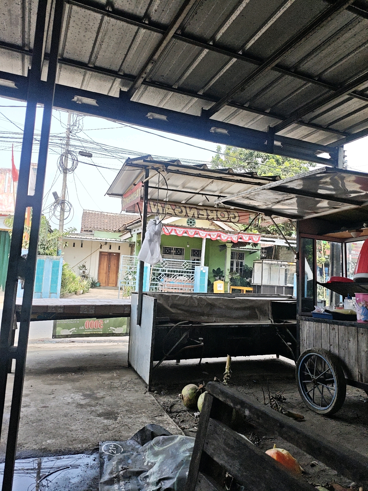 The image depicts an outdoor scene in Kemuning, Indonesia, likely in front of a food stall or small shop. A corrugated metal roof is prominent in the foreground, supported by dark metal beams. Below the roof, a green building with white trim and a red and white awning is visible. In front of this building, a portable stall with a wooden frame and a metal counter is present. A bicycle wheel is attached to the right side of this stall. The ground is a mix of concrete and dirt, with debris scattered around, including coconuts and what appears to be a pumpkin. Power lines are visible overhead, contributing to the urban environment. A red and white flag is also visible in the background on the left side of the image.