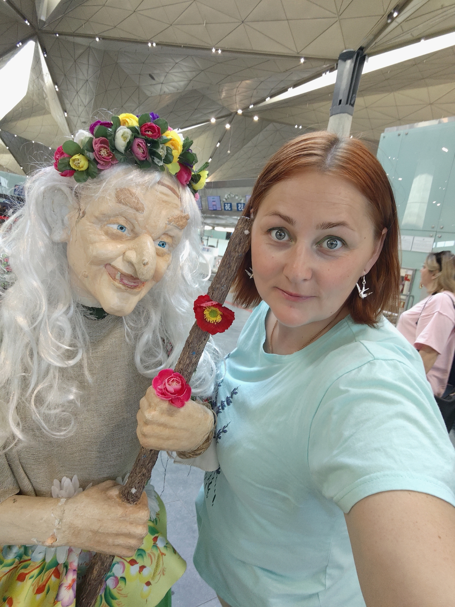 A woman with auburn hair and light blue eyes poses for a selfie with a figure of an elderly witch. The witch is wearing a floral wreath on her head, a rough-spun tunic, and a colorful skirt, and is holding a wooden staff adorned with red and pink flowers. The woman is wearing a light blue t-shirt and a necklace, and has bird-shaped earrings. The background shows the interior of a modern building with a high, geometric ceiling and metallic elements, suggestive of an airport terminal or convention center. A blurred figure of another woman in a pink shirt is visible in the background on the right. The scene appears to be located in Saint Petersburg, Russia, likely at the Pulkovo Airport.