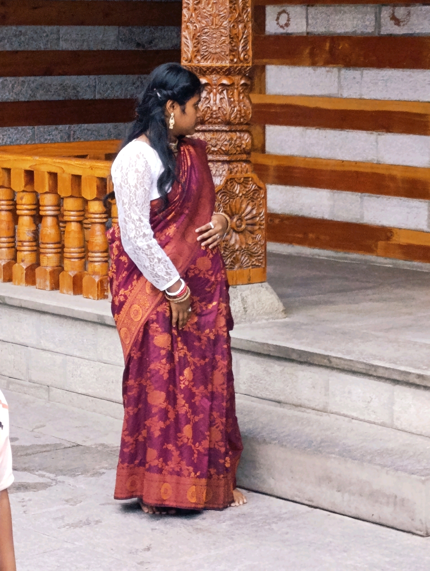 A woman stands with her back partially to the camera, wearing a deep maroon sari with gold floral patterns. She has a white lace blouse with long sleeves and her hair is styled in a braid with loose strands. She is adorned with gold earrings and multiple bangles on her right wrist. The woman appears to be standing on a stone staircase outside a building. To her left is a wooden railing with carved pillars. The background features a textured wall with horizontal wooden beams and stone blocks. The scene is outdoors, with natural light. This image was captured in Vashisht, India, likely near a temple or cultural site given the architectural style.