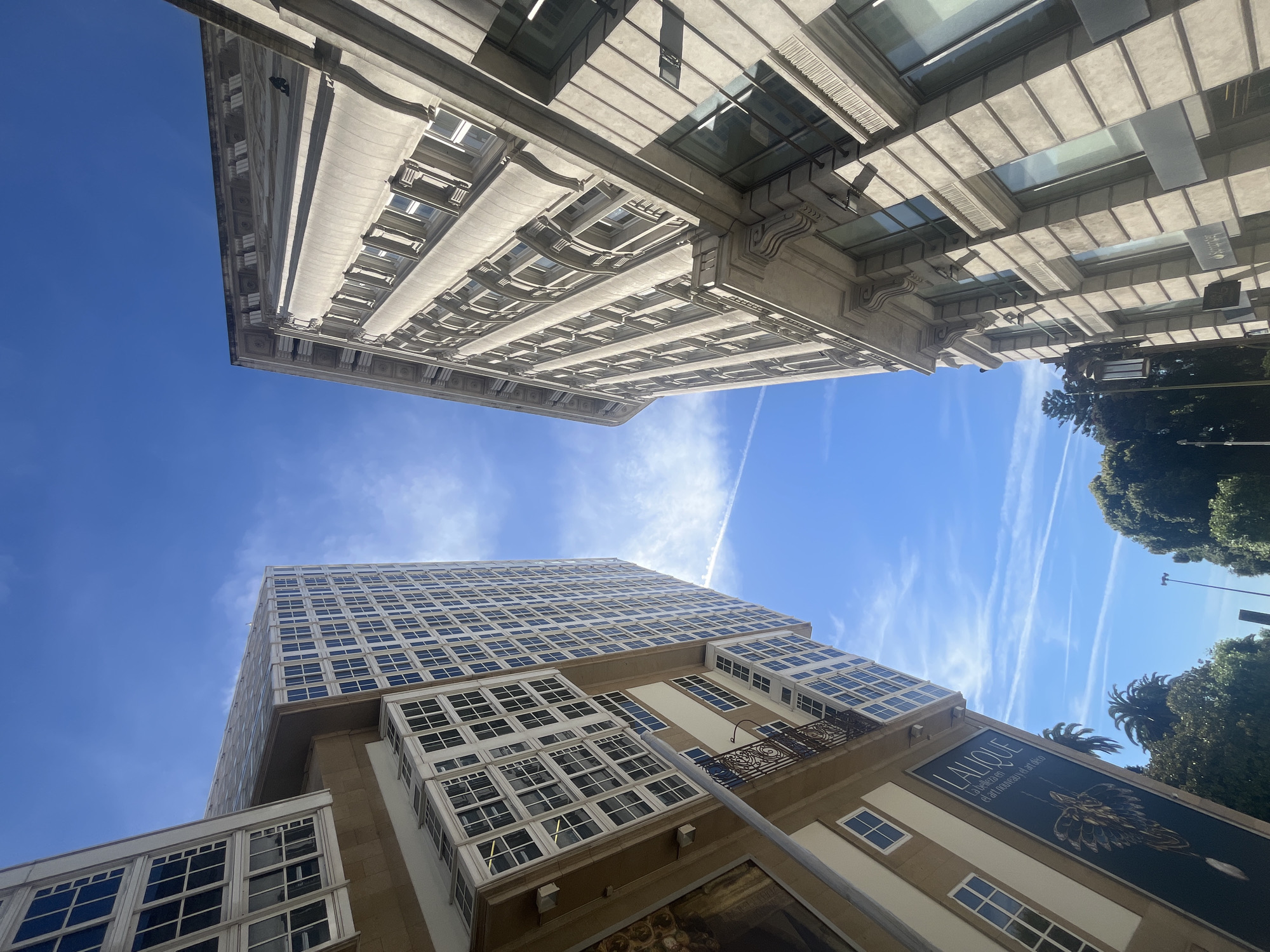 This is a low-angle, full shot of two buildings on a bright, sunny day in A Coruña, Spain. The building on the left is a modern high-rise with a glass facade and many windows, appearing to be residential or office space. It has a light-colored exterior. Below it, on the lower portion of the image, is another section of the building with a darker facade featuring a large poster or advertisement with the word "LALIQUE" and an ornate design. This section also has white-framed windows and a decorative black metal balcony railing.
The building on the right is a much older, ornate stone building with classical architectural details. It features columns, decorative cornices, and numerous rectangular windows. The stone appears to be a light grey or beige.
The sky is a vibrant blue with thin, wispy white clouds and several visible contrails from airplanes. Lush green trees, including palm trees, are visible at the base of the buildings and in the background. A lamppost is visible on the left side of the stone building. The perspective is tilted, making the buildings appear to lean towards the sky.