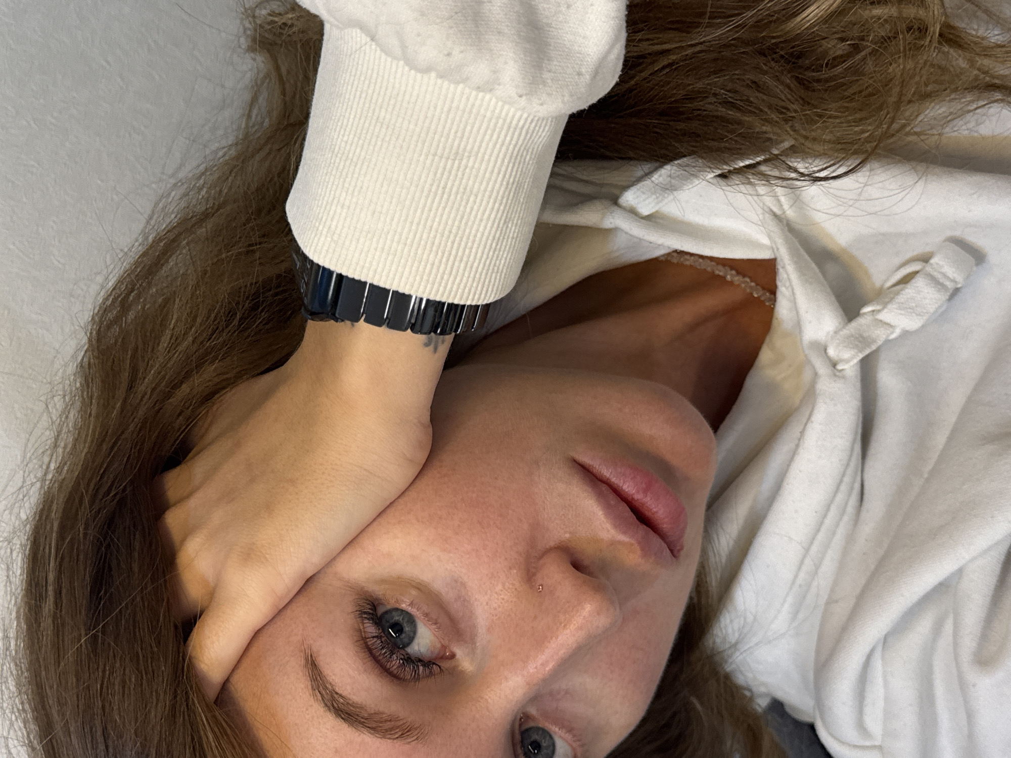 A woman with light brown hair and fair skin is lying down, angled towards the camera. She's wearing a white hooded sweatshirt. Her left hand is positioned near her face. She has blue eyes and a small piercing in her nostril. A black, metal watch is visible on her left wrist. The background appears to be a plain, light-colored wall. The image does not show the location of Chaikovsky, Russia.