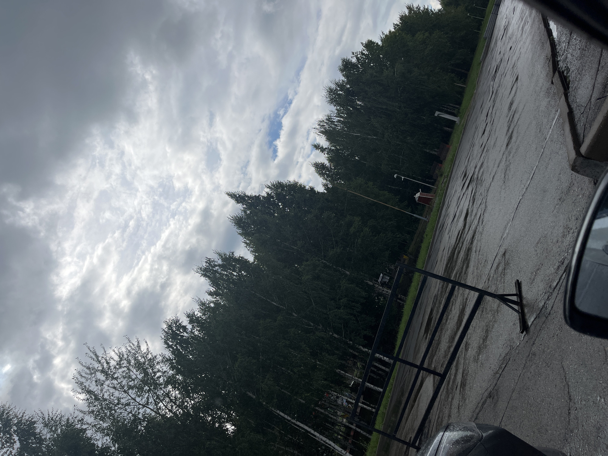 The image depicts a view of a paved area, possibly a parking lot, with a line of trees bordering it. The pavement appears wet, suggesting recent rain. A black metal barrier is set up in the foreground, angled across the paved surface. In the background, beyond the trees, there are glimpses of structures, including what might be lampposts and a small red building. The sky is cloudy. The scene is located in the city of Zainsk, Russia.