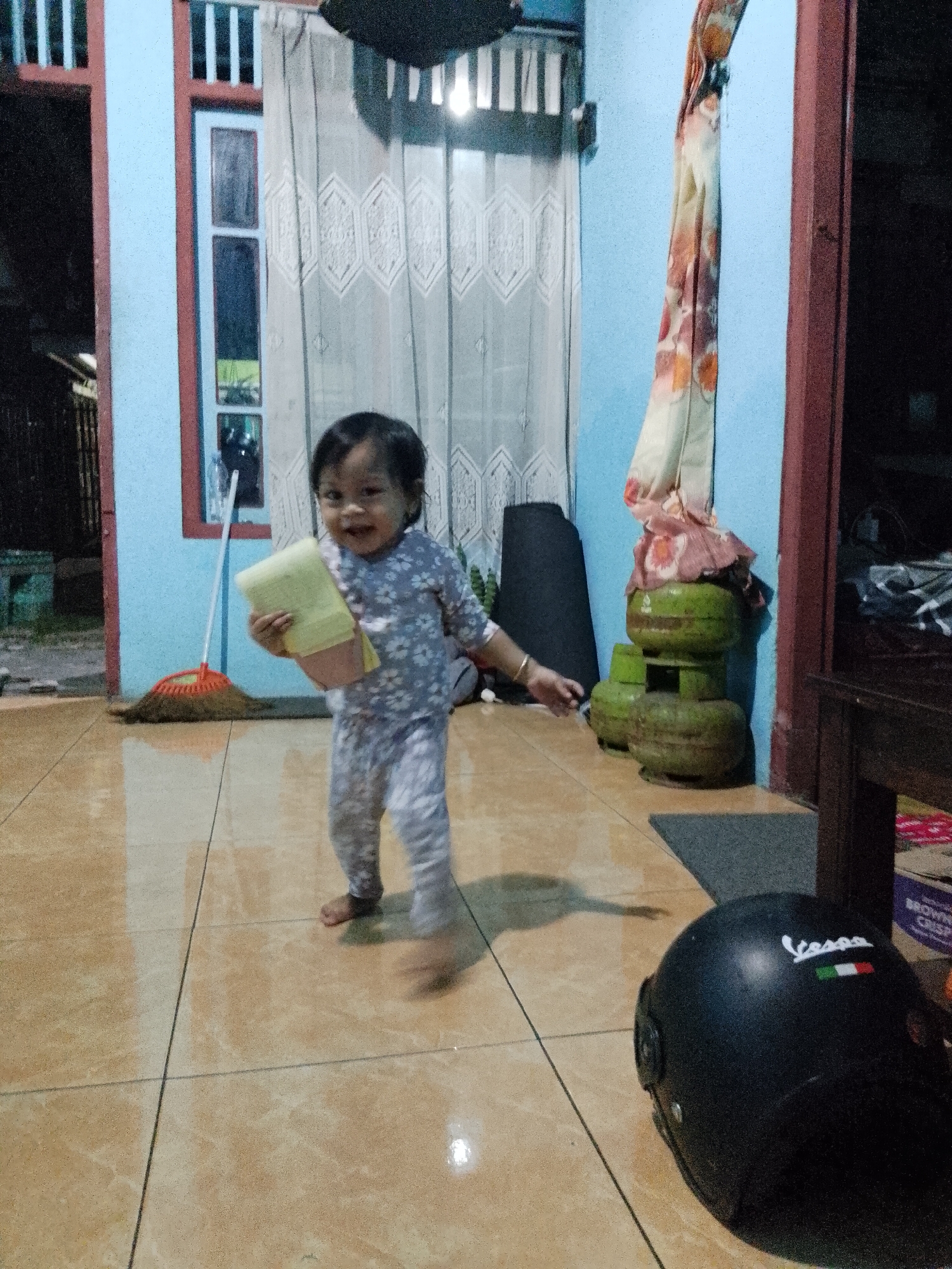 The image depicts a child, likely a toddler, standing in what appears to be an indoor residential setting, possibly a living room or hallway in Kecamatan Pati, Indonesia. The child is wearing a light-colored, patterned pajama set and is holding a yellow object, possibly a small towel or cloth, in their right hand. The child is smiling and looking towards the camera.
The floor is tiled with large, light brown tiles, and there are distinct grout lines. To the left of the child, a broom with an orange handle and a brown straw head is propped against a light blue wall. Behind the broom, a window with vertical white bars and red trim is visible, with patterned curtains hanging inside.
To the right of the child, three green gas cylinders are stacked vertically, with a floral patterned fabric draped over the top two. A dark gray rug is placed on the floor in front of the gas cylinders. Further to the right, a dark wooden table or cabinet is partially visible, and a black helmet with a white "Vespa" logo and the Italian flag is lying on the floor in the foreground. The lighting suggests it is either evening or nighttime, with artificial light sources illuminating the scene.