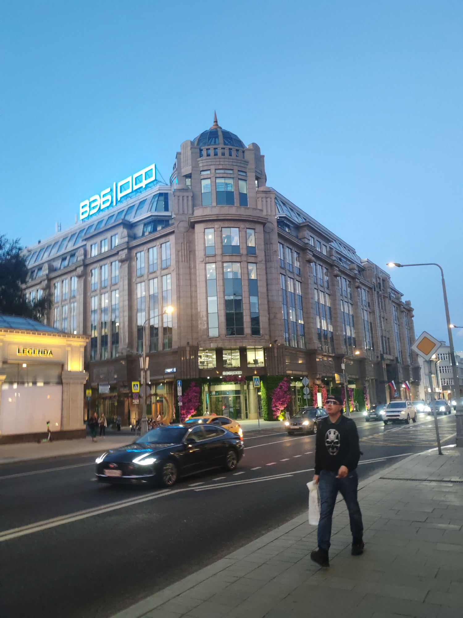 A man is walking on a sidewalk in an urban environment, with a multi-story building in the background and cars driving on the street. The building has a large neon sign with blue lettering on its roof and a dome on top. The man, wearing a black hooded sweatshirt with a skull graphic, blue jeans, and sunglasses, is holding a white bag in his right hand. The street is paved and has white lane markings. Several cars are visible on the street, including a black sedan in the foreground. The sky is a clear blue, indicating it is likely daytime or early evening. The sidewalk is made of stone tiles. The scene appears to be a commercial or business district.