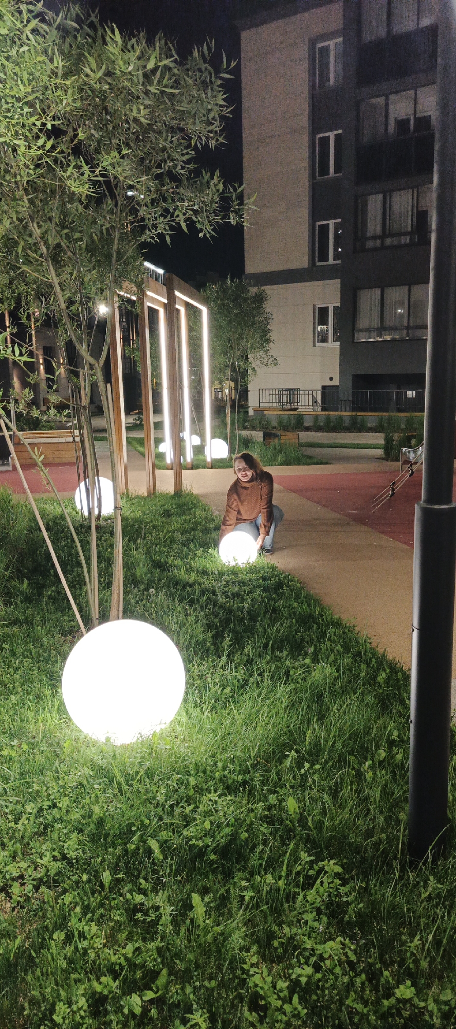 This nighttime image depicts an urban outdoor area in Izhevsk, Russia, likely a park or courtyard of a residential complex. A light-skinned female with light brown hair, wearing a brown ribbed turtleneck sweater and blue jeans, is positioned in a crouched stance on a light brown paved pathway, facing slightly towards the viewer. She is holding one of several glowing white spherical lights with both hands. Another identical glowing sphere is visible in the foreground to the left, resting within a patch of green grass.
The environment features multiple large patches of green grass interspersed with paved areas and sections of reddish-brown ground surfacing. In the midground, behind the individual, a series of tall, rectangular wooden structures with vertical light strips integrated into them are visible, forming an illuminated architectural element. Several slender trees with green foliage are scattered throughout the scene. In the background, modern multi-story apartment buildings are present; one section is light beige with white-framed windows, and an adjacent section is dark gray with larger windows and balconies. A dark cylindrical pole partially obscures the right side of the foreground. The overall lighting is artificial, emanating from the spherical lights, the vertical light strips, and the building windows, illuminating the immediate surroundings against a dark night sky.