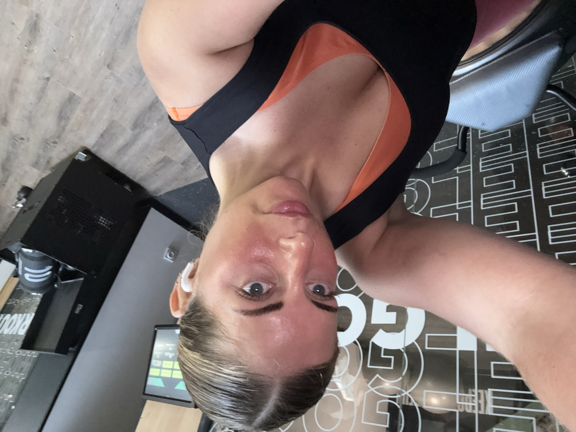 A selfie of a woman wearing a black and orange workout top and white earbuds. She is in a room with a black machine and a monitor on the left, and a patterned floor on the right. The location in the city of Paris, France is unknown.