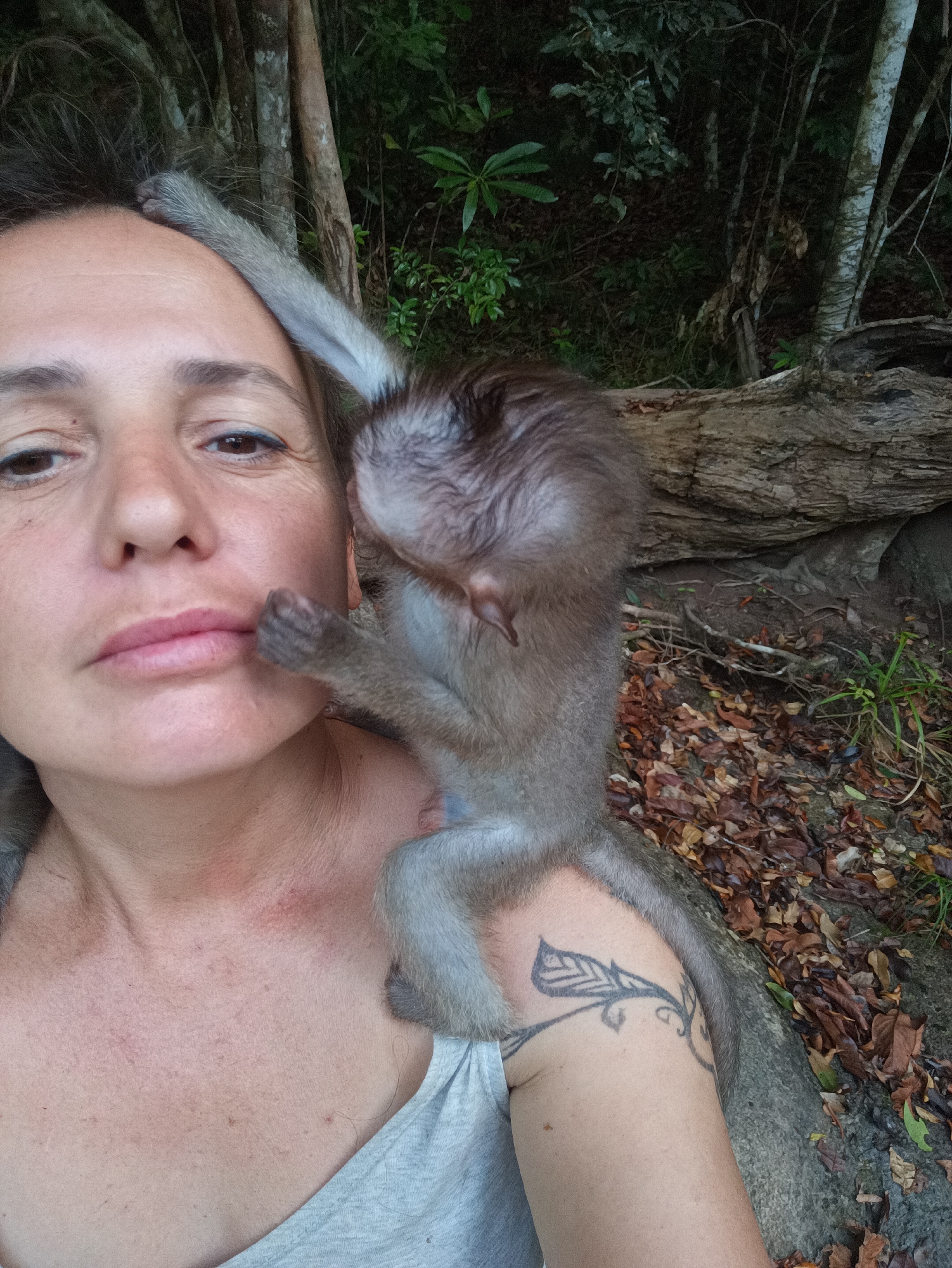 A woman poses for a selfie while a juvenile macaque monkey sits on her shoulder in what appears to be a forested area in or around Baan Thong Nang, Thailand. The woman is fair-skinned with dark hair, wearing a gray top. The monkey is gray-brown with small, human-like hands. The background includes trees, logs, leaves, and foliage.