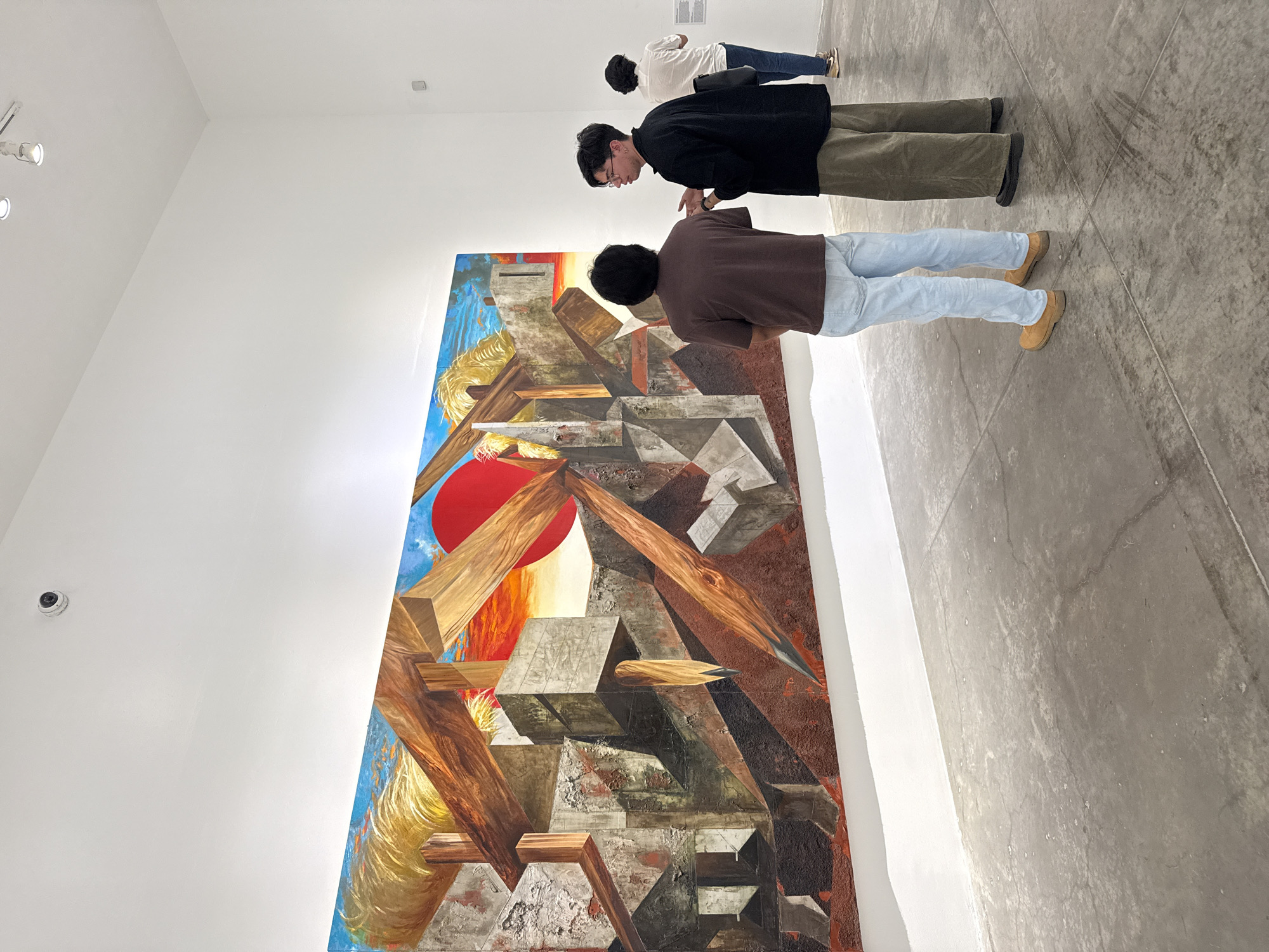 Three individuals are observing a large abstract painting in what appears to be an art gallery setting in Mexico City, Mexico. The painting, predominantly featuring reds, browns, and blues, is characterized by geometric shapes and linear elements. The three viewers, from left to right, are standing near the painting, their attention focused on the artwork. The gallery space is minimalistic with white walls and a concrete floor. A security camera is mounted on the wall near the artwork. The perspective of the photograph suggests it was taken from a slightly elevated angle.