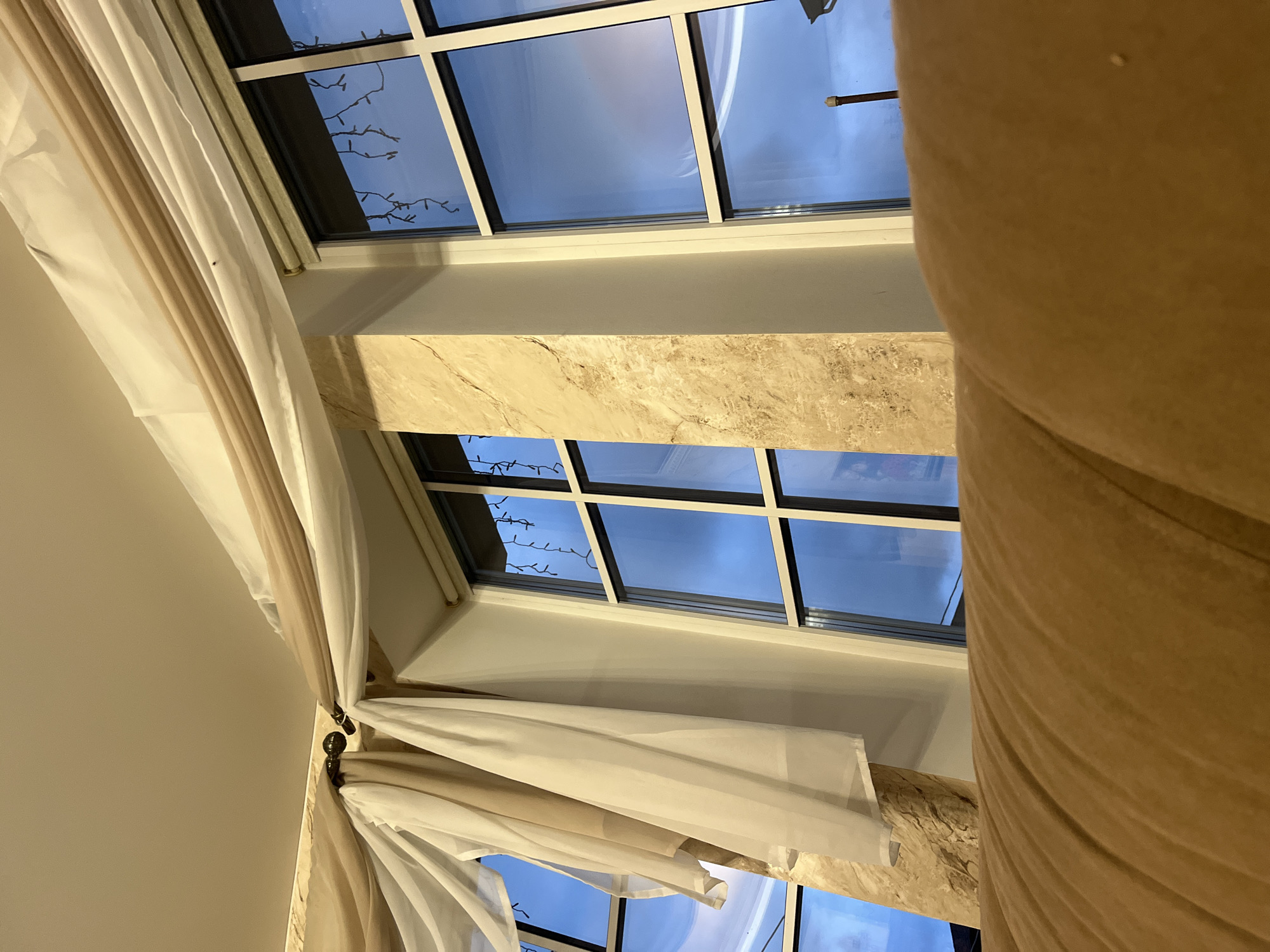 The image shows a close-up view of a window with sheer drapes in a residential setting, possibly a living room. The window has multiple panes and is decorated with string lights, suggesting a festive or decorative element. The drapes are a light, neutral color and are draped over a curtain rod. In the foreground, a portion of a large, cushioned piece of furniture, likely a sofa, is visible, rendered in a warm, earthy tone. The overall impression is one of an indoor domestic scene with an emphasis on window treatments and soft furnishings. The image does not contain any information indicating its location in Vladimirovka, Russia, or any people or actions.