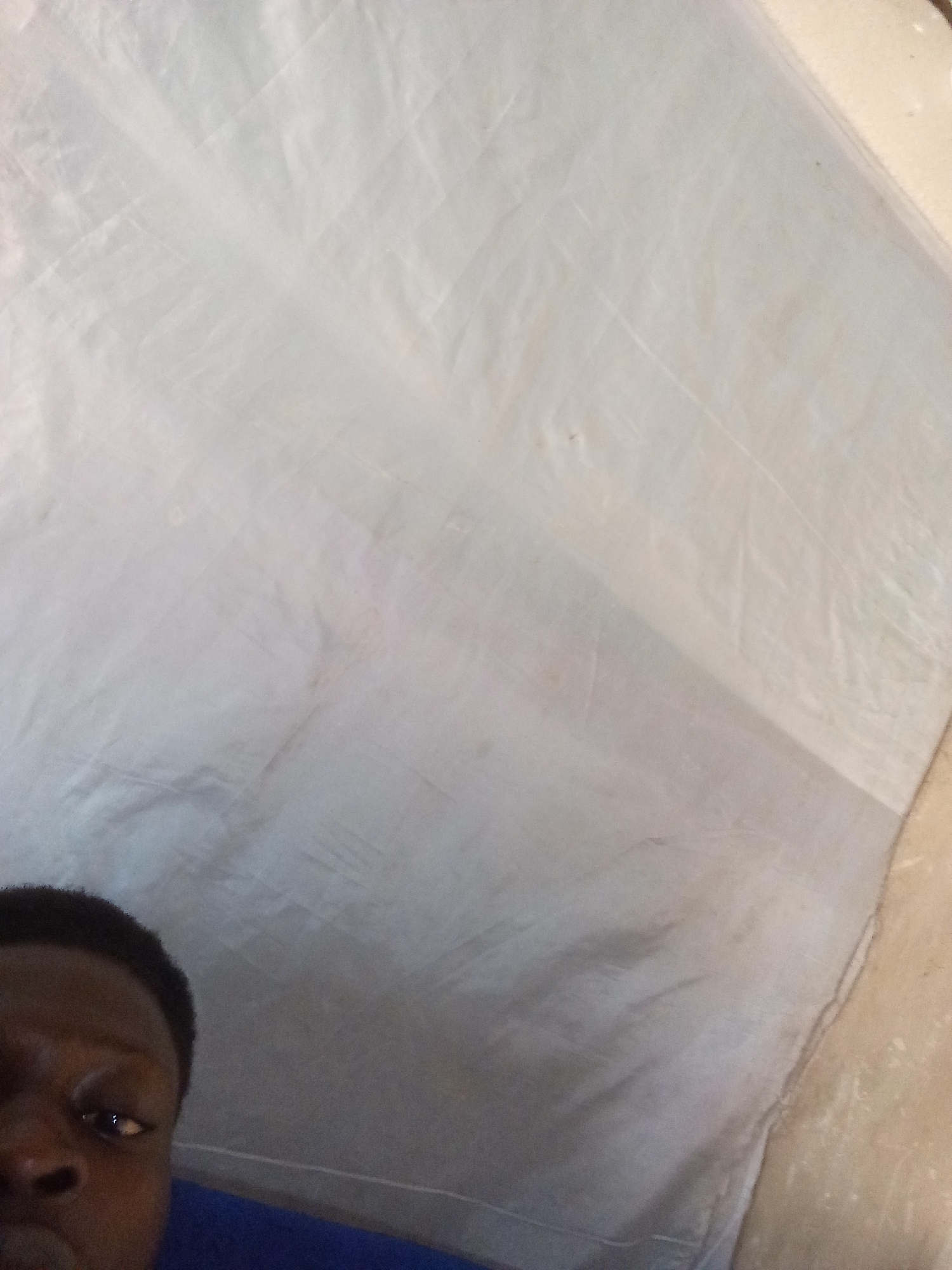 The image shows a close-up of a person's face and dark hair in the lower-left quadrant. The individual's left eye and part of their nose are visible. The rest of the image is dominated by a wrinkled, light-colored fabric that occupies the upper and right portions. The fabric appears to be draped or stretched, creating shadows and folds. The lighting is somewhat dim, with a noticeable gradient across the fabric, suggesting the light source is from the top right. The overall impression is that of a candid, indoor photograph, possibly taken with a mobile phone, with the background being a textile. The specific location in Nyabe, Nigeria, cannot be determined from the visual information provided in the image.
