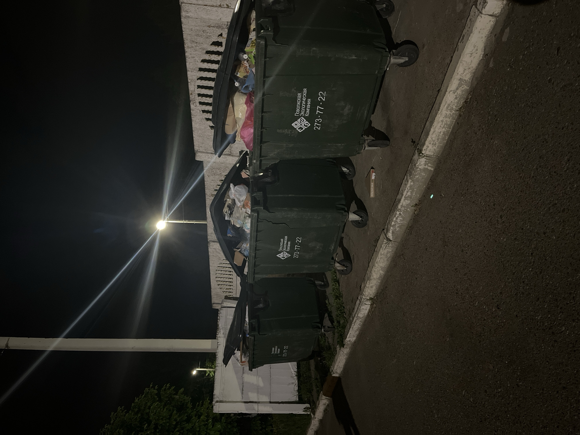 The media file presents an outdoor night scene featuring three large, dark green waste bins lined up alongside a concrete wall or fence. The bins are full, with their lids partially open, revealing various types of refuse, including plastic bags, cardboard, and other household waste. The scene is illuminated by a bright street light, visible in the upper left, which casts a strong beam across the dark sky.
Behind the bins, a textured concrete structure, possibly a retaining wall or barrier, is visible. To the right of the bins, there's an asphalt surface, likely a road or parking area, separated by a worn white curb. A small, reddish-white rectangular object, possibly a discarded cigarette pack or small box, lies on the ground near the base of the middle bin.
No people or active events are captured in the image, suggesting a quiet moment in an urban or suburban setting at night. The dim lighting, contrasted with the bright street light, creates a somewhat stark atmosphere. In the distant background, to the lower left, some dark foliage of trees or bushes can be discerned.
Visible text on the side of the waste bins, in Cyrillic, reads "Поволжская Экологическая Компания" (Volga Ecological Company) and a phone number "273-77-22". The small object on the ground has partially visible text including "LOVE". The setting, identified as Ореховка, Russia, aligns with the Cyrillic text on the bins.