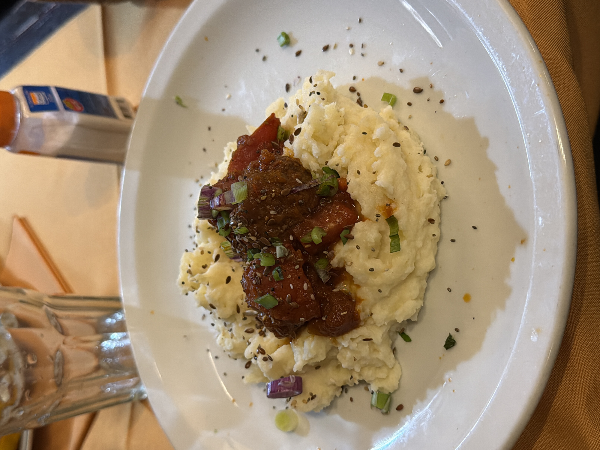 A top-down, medium shot shows a white plate with a serving of mashed potatoes and what appears to be braised short ribs. The dish is garnished with sliced green onions and seeds. The plate is resting on a brown tablecloth. A bottle of seasoning is visible to the left of the plate, and a glass is also visible. The image does not contain enough information to establish a location in the city of Buenos Aires, Argentina.