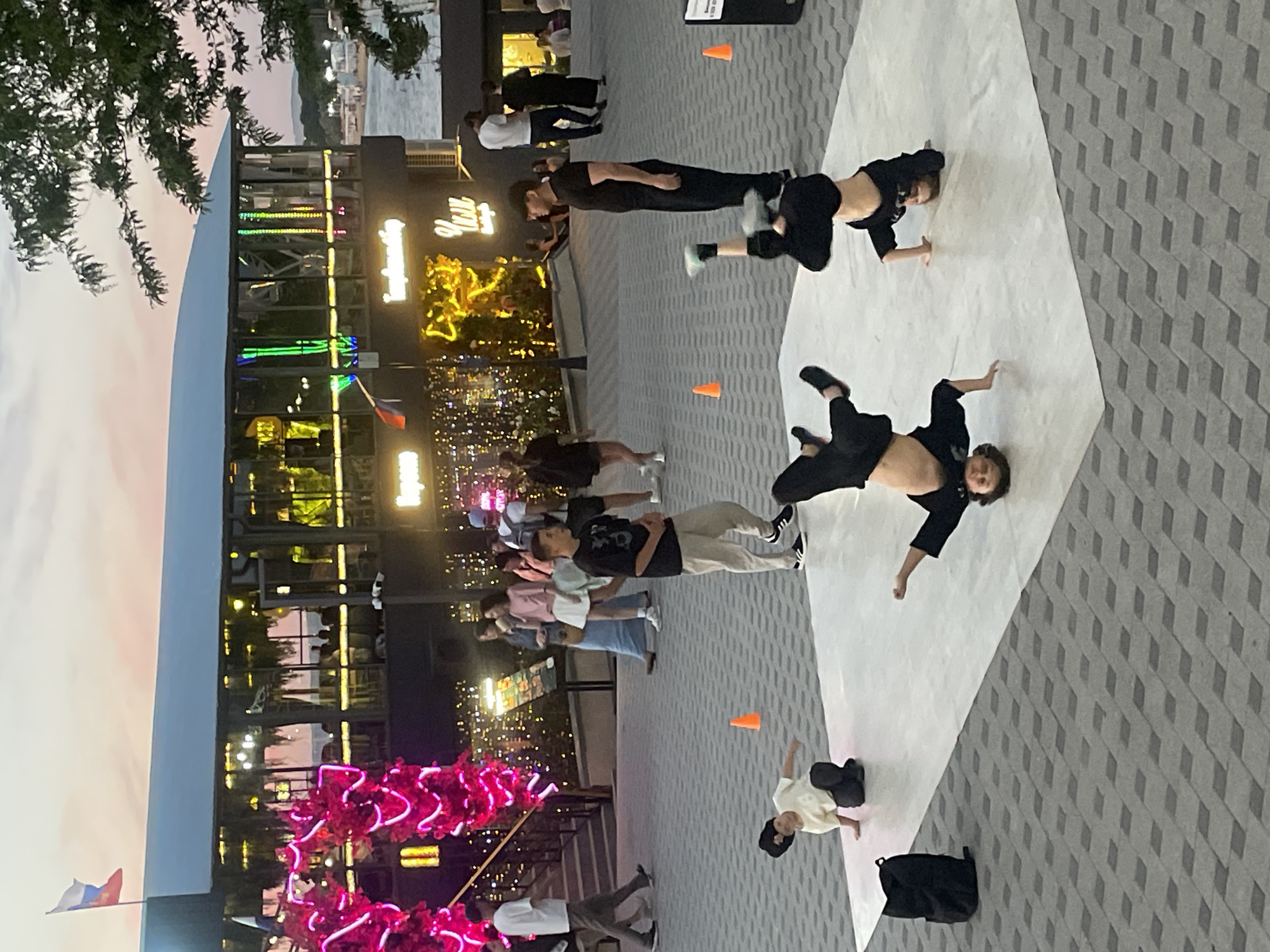 The image depicts an outdoor evening scene in Anapa, Russia, likely on a waterfront promenade. In the foreground, four individuals are engaged in breakdancing on a white mat laid out on a patterned stone surface. Three dancers are upside down, performing power moves, while one is on their knees. To the left of the mat, a black backpack rests on the ground. Several orange traffic cones are visible around the mat.
In the background, a modern building with a glass facade is illuminated, suggesting it might be a restaurant or entertainment venue. The building is adorned with bright pink neon decorations on its left side and string lights with warm yellow bulbs illuminating the area around it. People are scattered throughout the background, some walking and others observing the breakdancing performance. A Russian flag flies from a pole on the roof of the building. The sky above is a soft gradient of pink and blue, indicating dusk. The overall atmosphere suggests a lively public space with recreational activities.