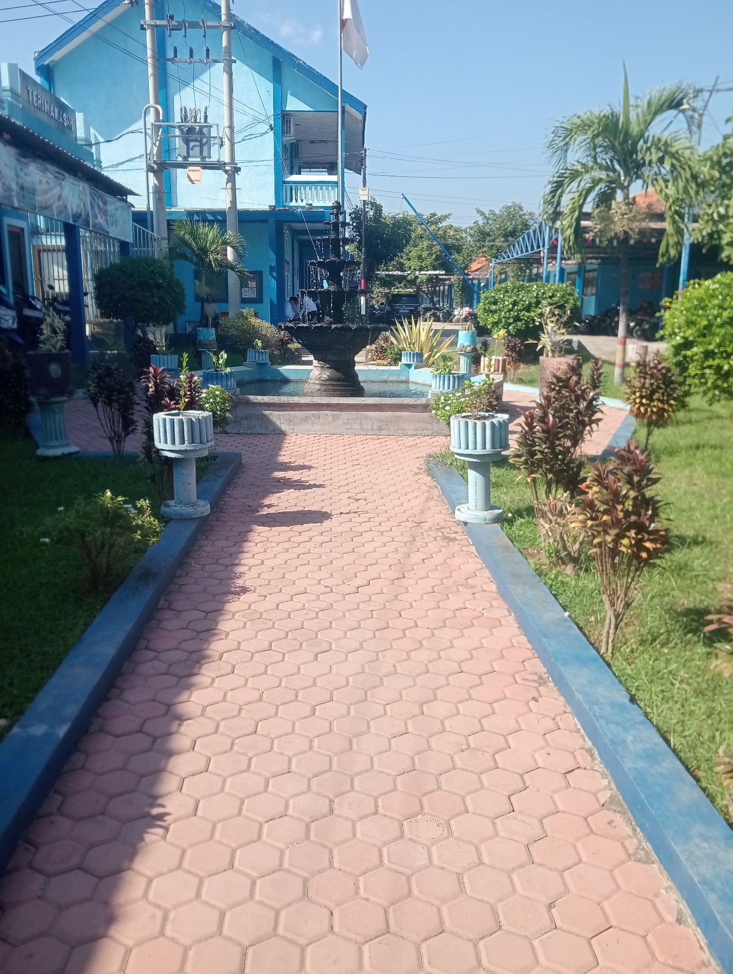 The image shows a view of a building with blue walls and a tiled pathway in the city of Buduan, Indonesia. The building appears to be a school or administrative complex, with visible signs and electrical equipment. The pathway is made of hexagonal orange tiles and is bordered by blue concrete. Several potted plants and trees are scattered throughout the scene, and a fountain is visible in the background. The sky is clear and blue, indicating a sunny day. There are no people clearly visible in the image.