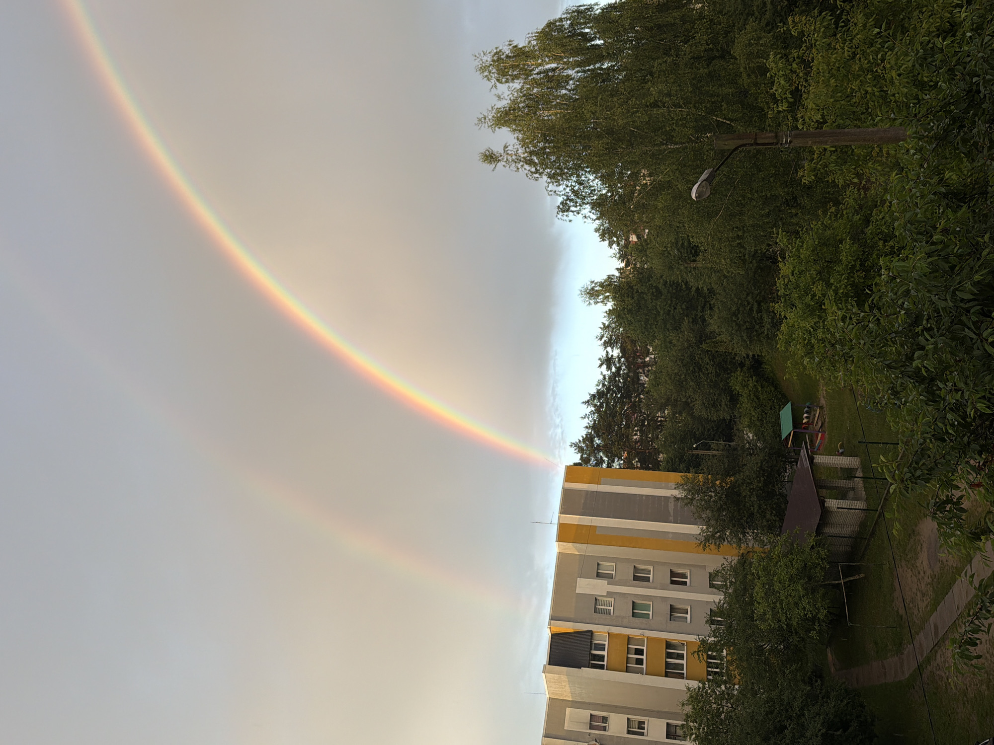A double rainbow arches across a cloudy sky above a residential area in Novoiavorivsk, Ukraine. The primary rainbow, with its full spectrum of colors, is prominent, while a fainter secondary rainbow is visible above it. Below the sky, a multi-story apartment building with a grey and yellow facade stands to the left. The building has numerous windows and balconies. Dense green trees and foliage surround the building and extend to the right of the frame. A small, open-air shelter with a dark roof and brick supports is situated in a grassy courtyard, adjacent to a children's play area with a sandbox. A street lamp with a curved arm is visible amongst the trees on the right side of the image. The ground in the foreground shows a paved pathway and grass.