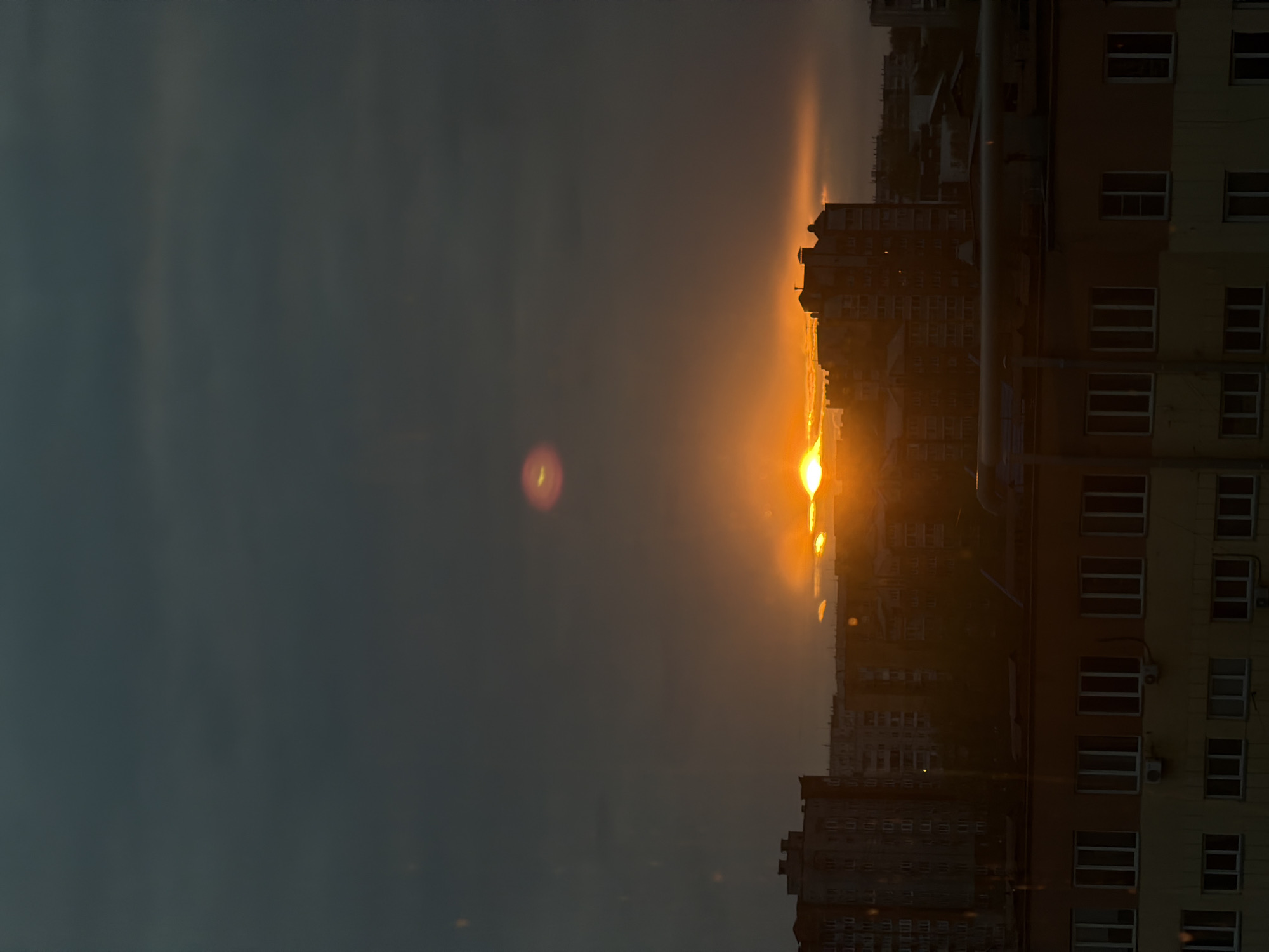 The image depicts a cityscape at sunset, likely taken from a high vantage point in Krasnoyarsk, Russia. The sun is setting behind a cluster of buildings, casting a warm, golden light on the surrounding structures. The sky is a mix of dark grey and orange hues, reflecting the setting sun. In the foreground, there is a residential building with multiple windows, some of which appear to have air conditioning units installed. The overall impression is that of a peaceful evening in an urban environment.