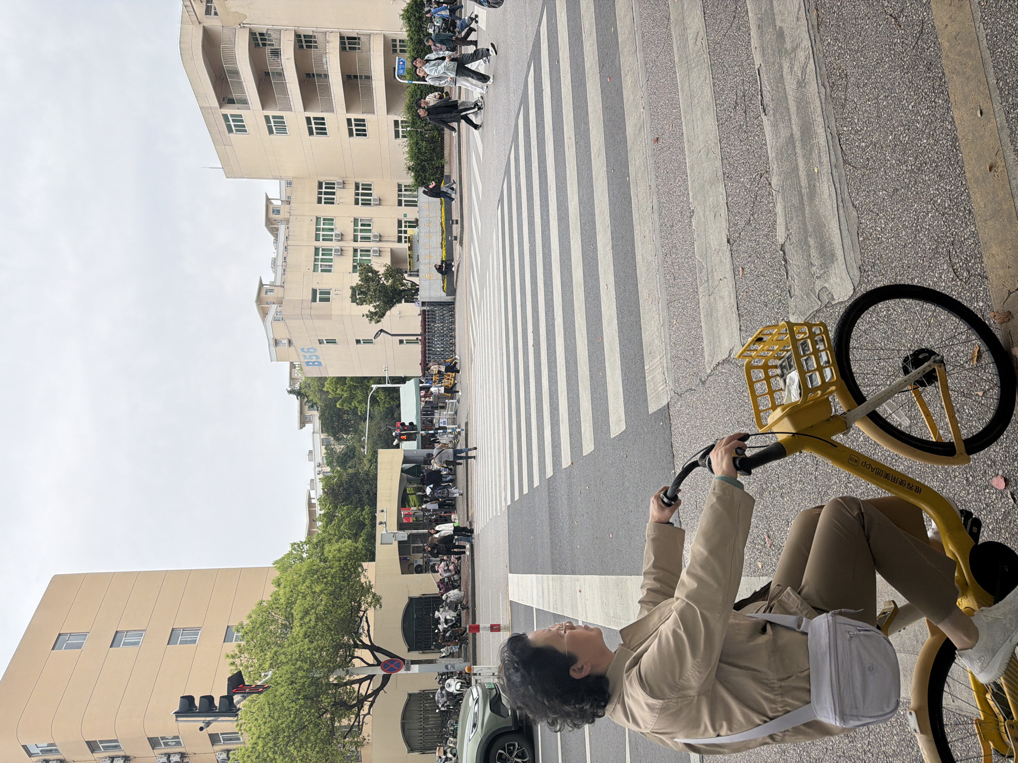 A person is riding a yellow bicycle on a zebra crossing in Jiangning District, China. The person is wearing a beige jacket, brown pants, and white shoes. They also have a small white bag over their shoulder. Buildings are in the background, including a light-colored building on the left with several stories and windows, and other buildings further down the street. Pedestrians are crossing in the background. A car and several motorbikes are parked by the side of the road. The sky is overcast.