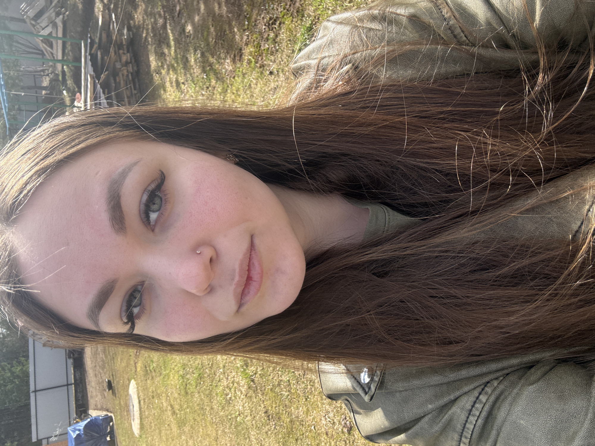 A selfie of a young woman with long brown hair and fair skin is taken outdoors in daylight. She is wearing a green jacket. Her left nostril has a piercing. The background includes grass, trees, and some indistinct structures, suggesting a park or rural setting. There is no specific information to confirm that the location is in Ольховка, Russia.