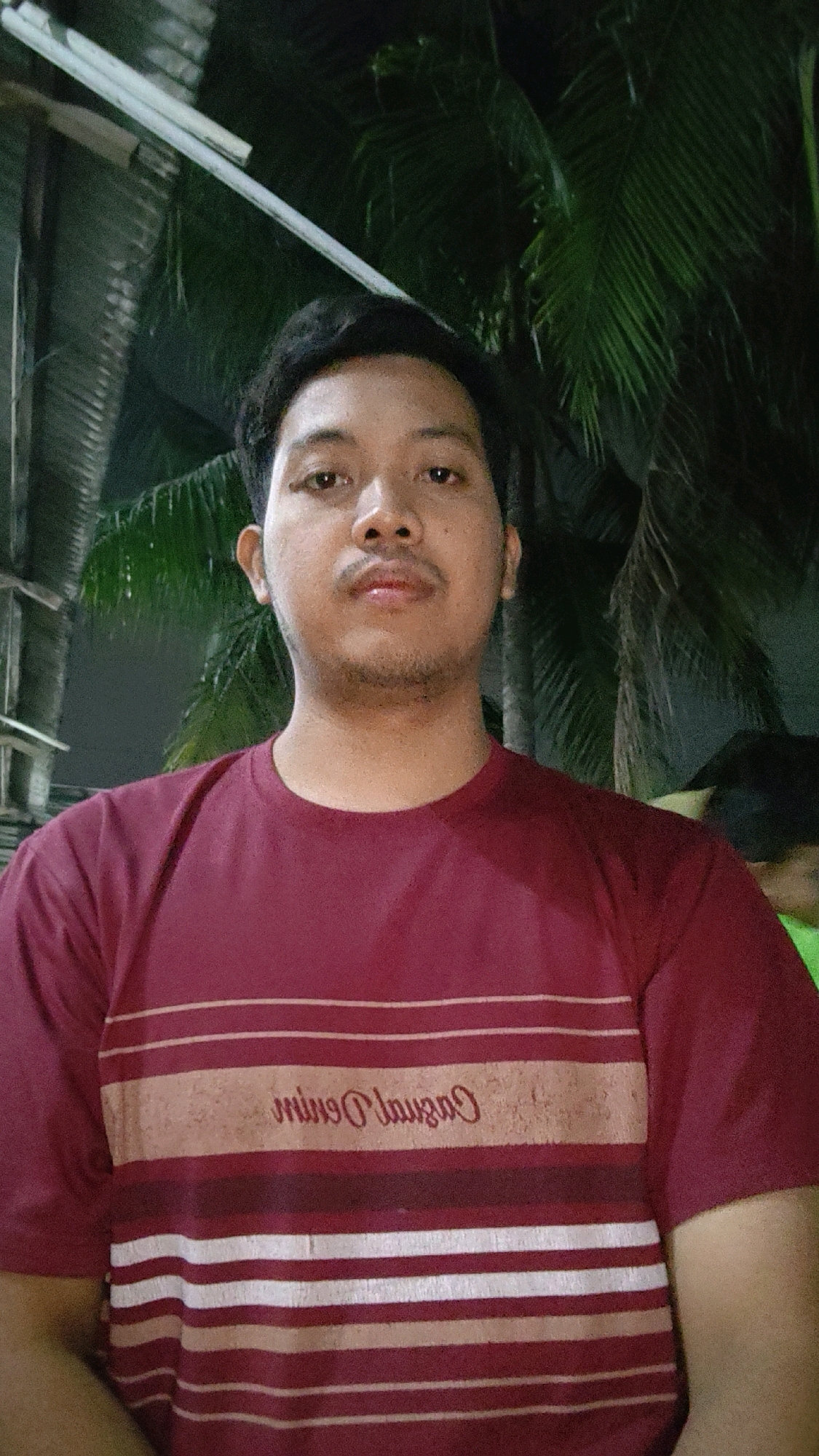 A young man with dark hair and a light complexion is photographed from the chest up. He is wearing a maroon t-shirt with horizontal stripes in varying shades of red, cream, and white. A logo is visible on the t-shirt, in cursive script, reading "Casual Denim." He has a mustache and a hint of stubble on his chin and jawline. The background is dark, with the silhouette of palm tree fronds and some corrugated metal roofing visible. The lighting suggests it is nighttime. The image was taken in Mekarmukti, Indonesia.