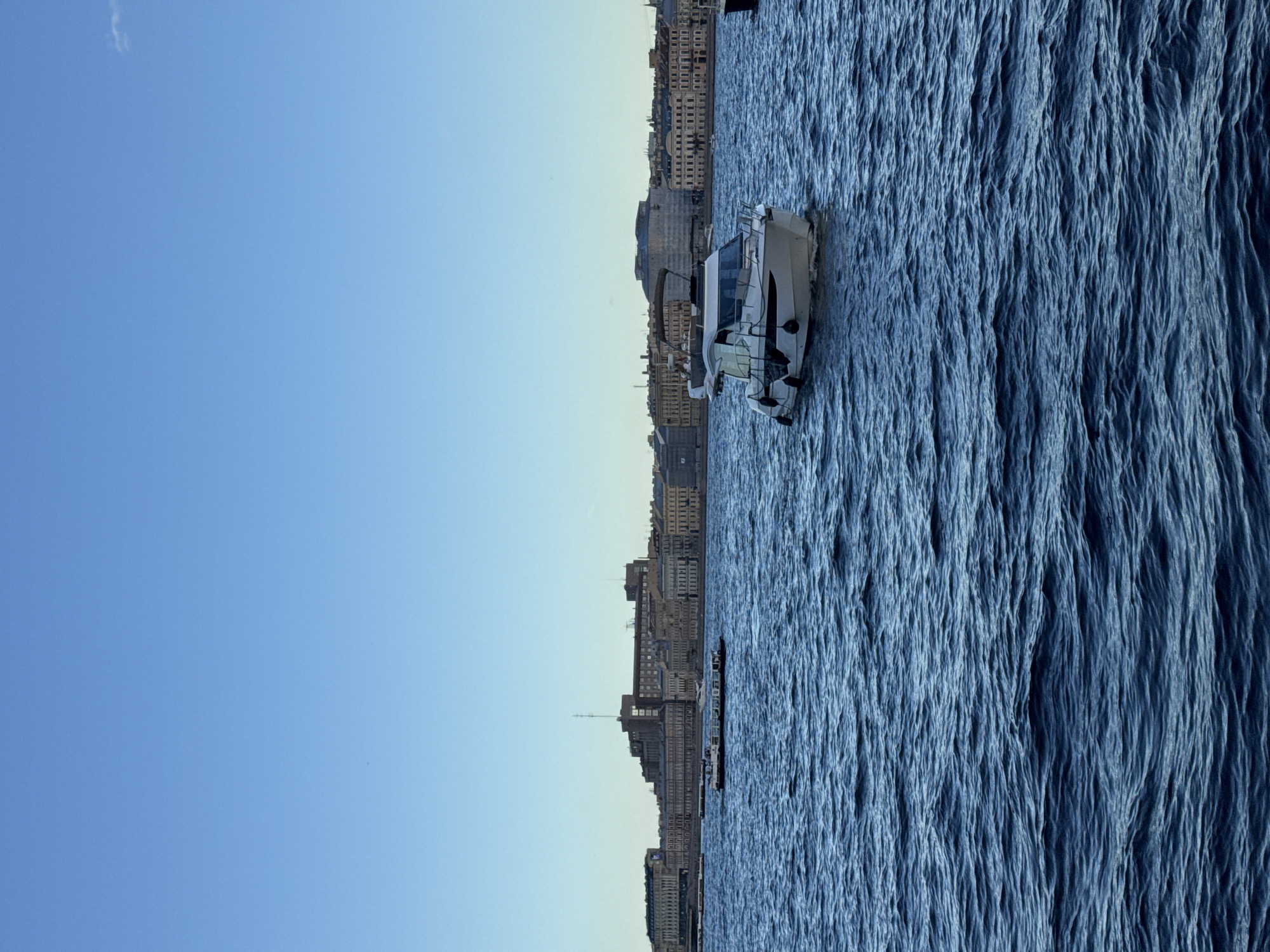The image depicts a scene on the Neva River in Saint Petersburg, Russia, with the city's historic embankment visible in the background under a clear blue sky. In the foreground, the deep blue water of the river is rippled by a gentle breeze. A white motor yacht, the primary subject, is moving away from the viewer towards the right side of the frame. It features a closed cabin, a railing around the bow, and several black fenders hanging along its side. In the middle ground, a larger, longer white vessel, possibly a tour boat, is traveling parallel to the shore. The Neva River embankment is lined with classical architecture, characterized by its light-colored facades, numerous windows, and ornate details. Dominating the center of the embankment is a large, imposing brick building with a tall antenna structure on its roof, likely a significant administrative or institutional building. The overall impression is one of a serene day in a grand European city.