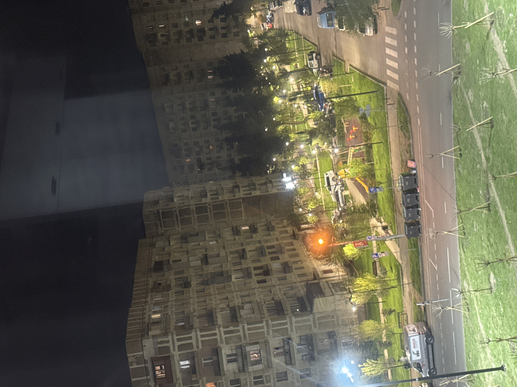 This nighttime image, rotated 90 degrees clockwise, captures an urban residential scene in Tbilisi, Georgia, from an elevated perspective. The dark sky indicates the time of day, with artificial light sources illuminating the area.
The scene features two large, multi-story apartment buildings with numerous windows, many of which show interior lights. Their architectural style appears characteristic of Brutalist or post-Soviet construction.
A paved road, marked with white dashed and solid lines, runs through the center, complete with a pedestrian crossing marked by zebra stripes. Several vehicles are visible, including parked cars and a white delivery van bearing the text "RICO" on its side. A blue van and a truck are also discernible.
Alongside the road lies a green park area, lush with grass and trees. Both mature trees and younger saplings, supported by wooden stakes, populate the space. The park is brightly lit by multiple streetlights, one emitting a distinct orange glow. Benches and a children's playground with visible slides and swings are present within the park. Street signs, including one indicating "P" for parking, are also scattered throughout the area. The image portrays a quiet, well-lit urban environment after dark.