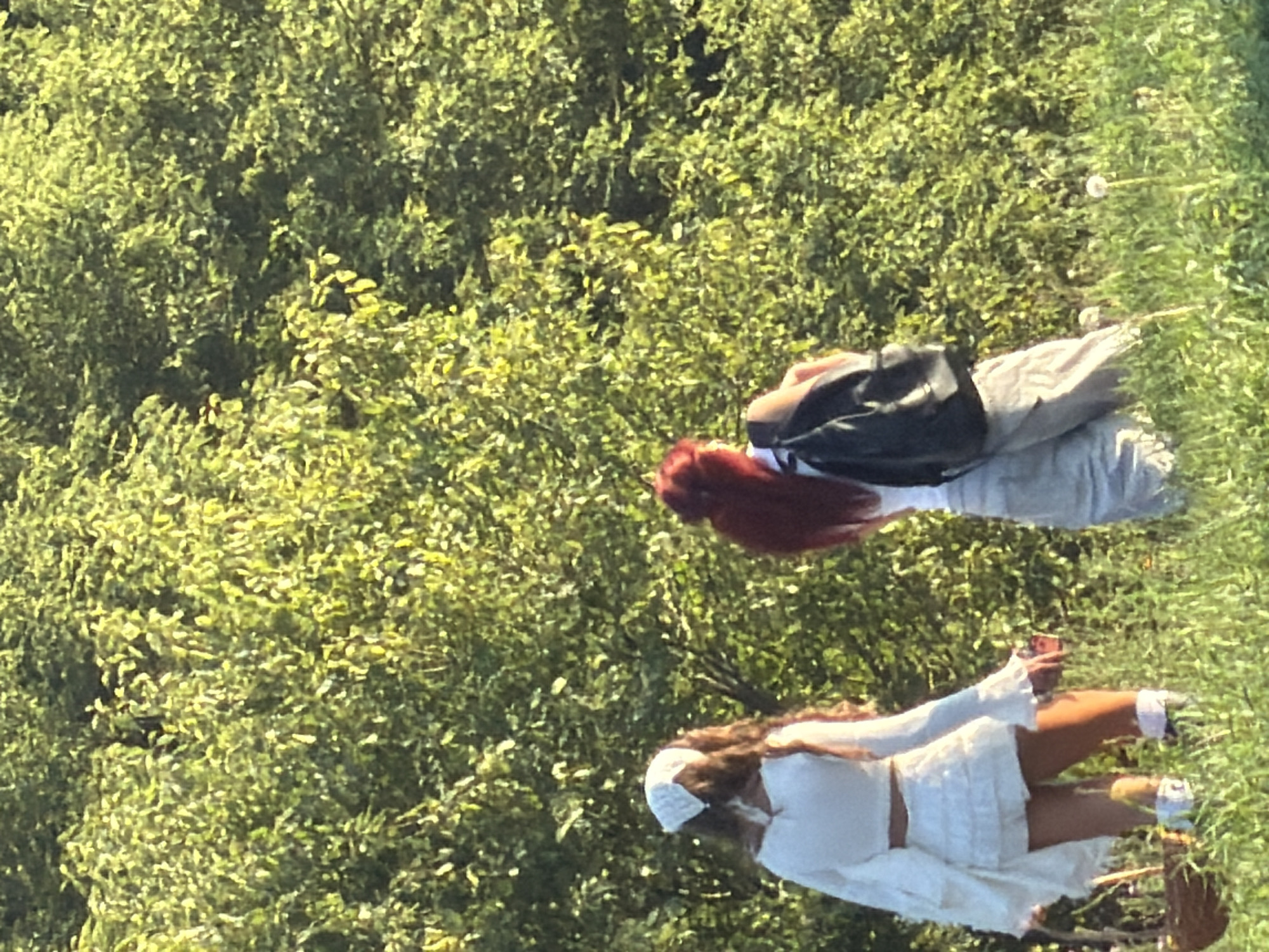 In Скорняково, Russia, two individuals are captured from behind, standing in a grassy area with a dense green foliage backdrop. The person on the left wears a white outfit consisting of a cropped top with bell sleeves and a ruffled skirt, accessorized with a white headscarf. They are holding a woven basket and a small object in their hands. The person on the right has bright red hair and is wearing a white top and light-colored pants, carrying a black backpack. The environment suggests an outdoor setting, possibly a park or a field, under natural lighting.