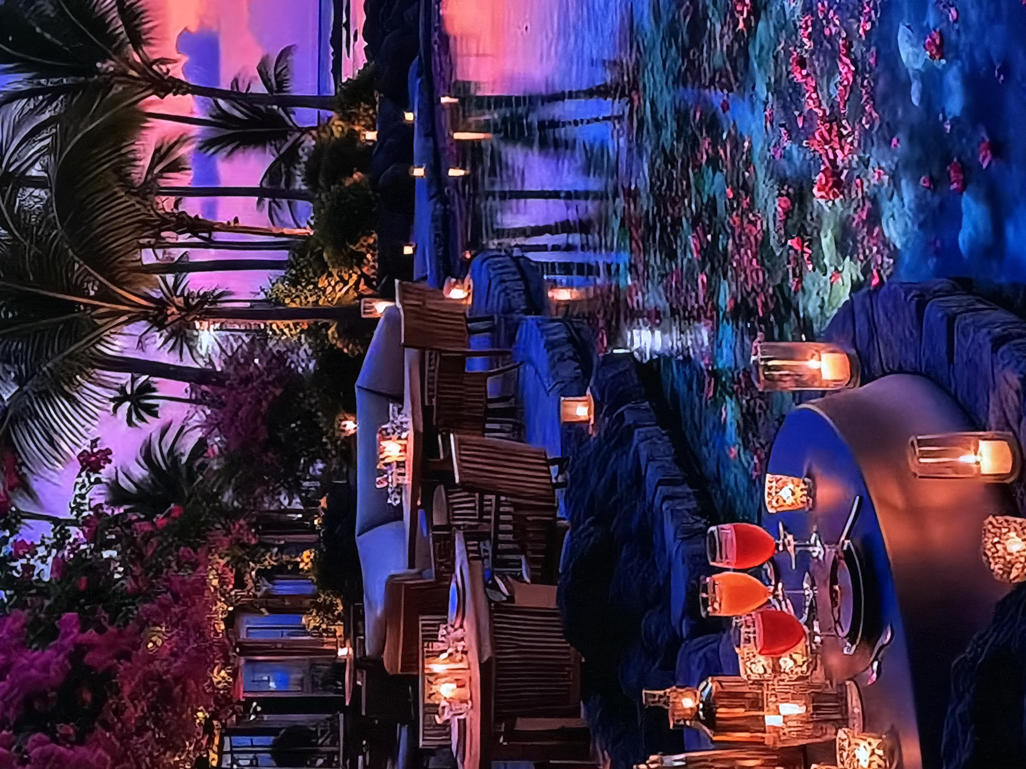 This image captures a vibrant, tropical-themed dining and lounge area, likely an upscale restaurant or event space, in Sofiivska Borshchahivka, Ukraine. The scene is set at what appears to be twilight or night, characterized by rich, artificial lighting in hues of deep purple, pink, and blue, creating a romantic and somewhat surreal atmosphere.
The foreground and left side of the frame feature a series of elegantly arranged tables and chairs, suggesting an outdoor dining experience. The chairs are made of slatted wood, and the tables are adorned with numerous candles in glass holders, emitting a warm, inviting glow. On one table in the foreground, three wine glasses holding drinks of vibrant red and orange liquid are visible.
Lush tropical vegetation, including tall palm trees with their distinct fronds, rises majestically on the left, silhouetted against the colorful backdrop. Interspersed among the palm trees are abundant bushes with bright pink and red flowers, possibly bougainvillea, adding splashes of vivid color to the scene.
A calm body of water runs through the right side of the image, beautifully reflecting the purplish-blue ambient light and the surrounding foliage. Red floral elements or petals are scattered across or floating on the water, enhancing the picturesque and serene setting. The combination of exotic flora, soft candlelight, and dramatic colored lighting creates a luxurious and tranquil ambiance, designed for an immersive and relaxing experience. The absence of people suggests a quiet moment, perhaps before opening or during a lull. Given the location in Ukraine, this tropical environment is almost certainly an elaborately designed indoor space. No text is visible in the image.