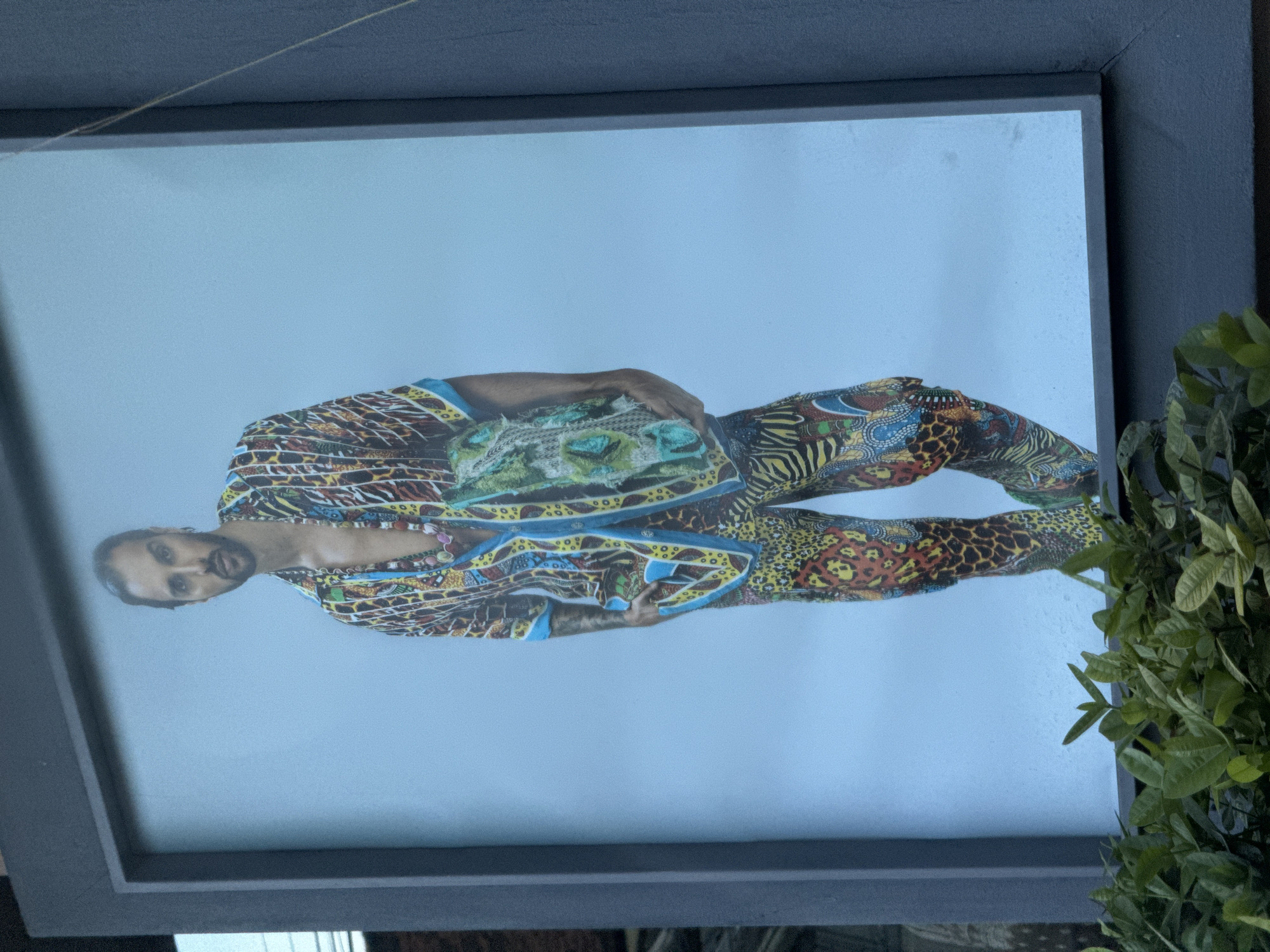 The media file displays a framed picture, rotated 90 degrees clockwise, making the framed subject appear horizontal. The dark gray/black frame is partially obscured on the right side by green foliage, possibly a plant.
Within the frame, a person with dark hair and a beard is depicted against a plain, light blue background. The individual is wearing a strikingly colorful and intricately patterned outfit, resembling a jumpsuit or a matching set. The attire features a vibrant mix of animal prints, including leopard spots and zebra-like stripes, combined with geometric and abstract designs. Dominant colors in the pattern are yellow, blue, red, green, and black. The person is looking directly at the viewer with a neutral expression. Their arms are bent, and they are holding a textured, possibly beaded or embroidered, object against their midsection, which also features patterns complementing the outfit. A necklace is visible around their neck.
No specific activity is shown beyond posing for the photograph. There are no visible cues within the image itself to indicate the time of day, weather, or location, though the provided context places the image capture in North Kuta, Indonesia. There is no visible text in the image.