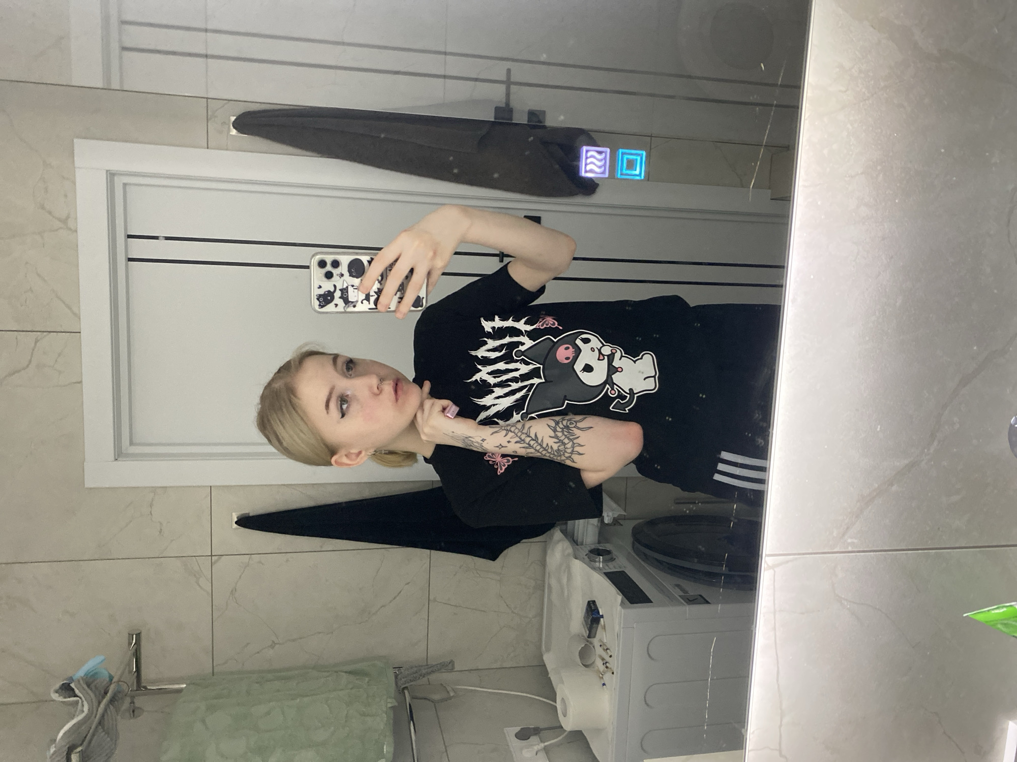 A young woman is taking a selfie in a bathroom. She is holding a phone with a white case adorned with black and white cat drawings. She has blonde hair and is wearing a black t-shirt with an anime character printed on it and black pants with white stripes. She has tattoos on her left forearm.
The bathroom features light-colored, marble-patterned tiles on the walls and floor. There is a mirror, a towel hanging above the door, a washbasin with a green sponge, a toilet paper holder, and a washing machine in the scene.
No information within the image suggests the location is Izhevsk, Russia.