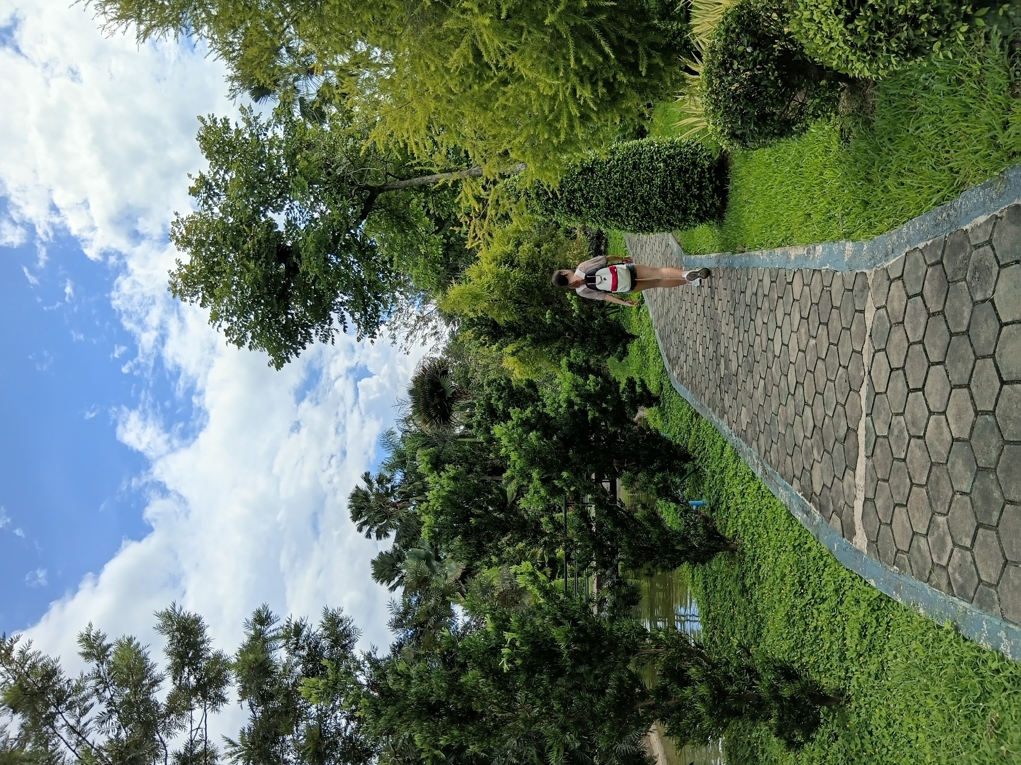 A person walks along a paved path in a park in Ban Pa Phai Si Khong, Thailand. The path is made of hexagonal gray bricks with a blue border. The person is seen from behind, wearing a light-colored shirt, shorts, and a backpack with white and red accents. They are walking away from the camera towards a wooded area. The park features lush green grass, various types of trees, and a body of water on the left side of the path. The sky is blue with scattered white clouds.