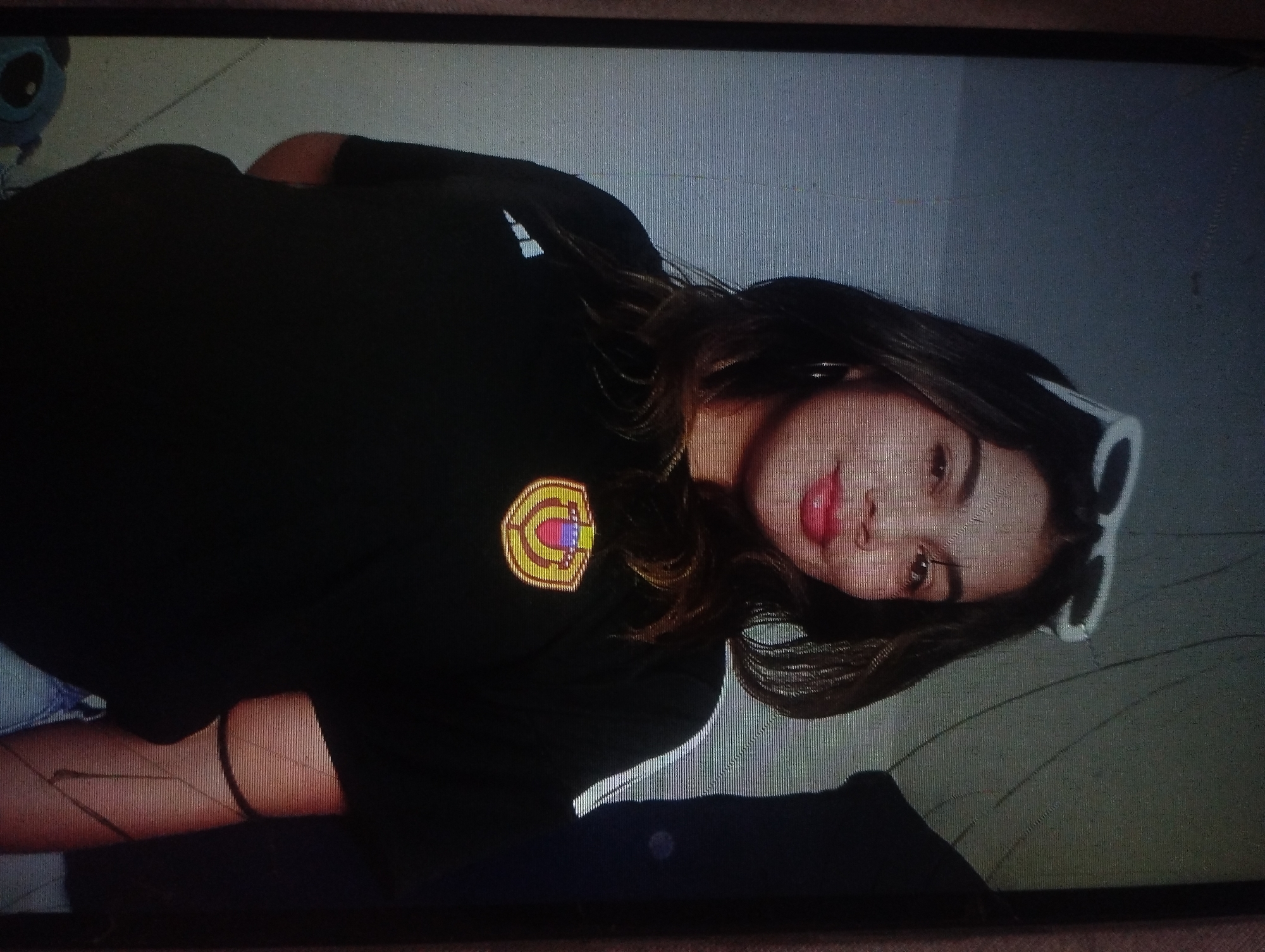 A young woman with long brown hair is lying down, turned to her left, and facing the camera. She is wearing a black t-shirt with a round logo on the left breast, white-framed sunglasses perched on her head, and a thin black bracelet on her left wrist. The background appears to be a neutral color, and a Stitch plushie is visible in the upper left corner. There is no visual information provided to determine the location in the city of Punta de Piedras, Venezuela.