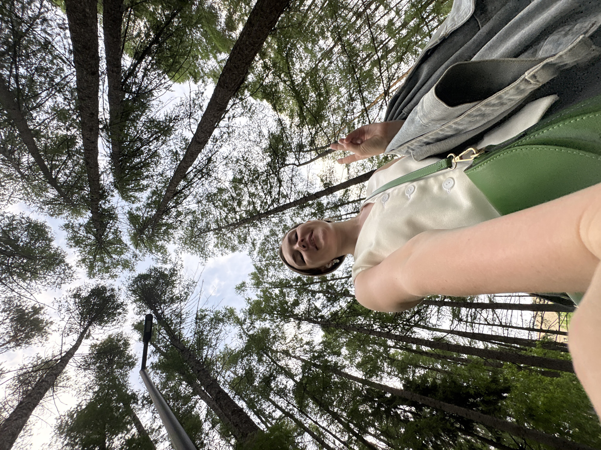 A low-angle, tilted, outdoor shot shows a person and the sky with trees. The person has fair skin, light brown hair, and a white top. Part of a denim jacket is visible, along with a green bag with a gold clasp. The background consists of tall trees reaching up to the sky. A street light is also visible. It is not possible to confirm the exact location in Izhevsk, Russia based on the image.
