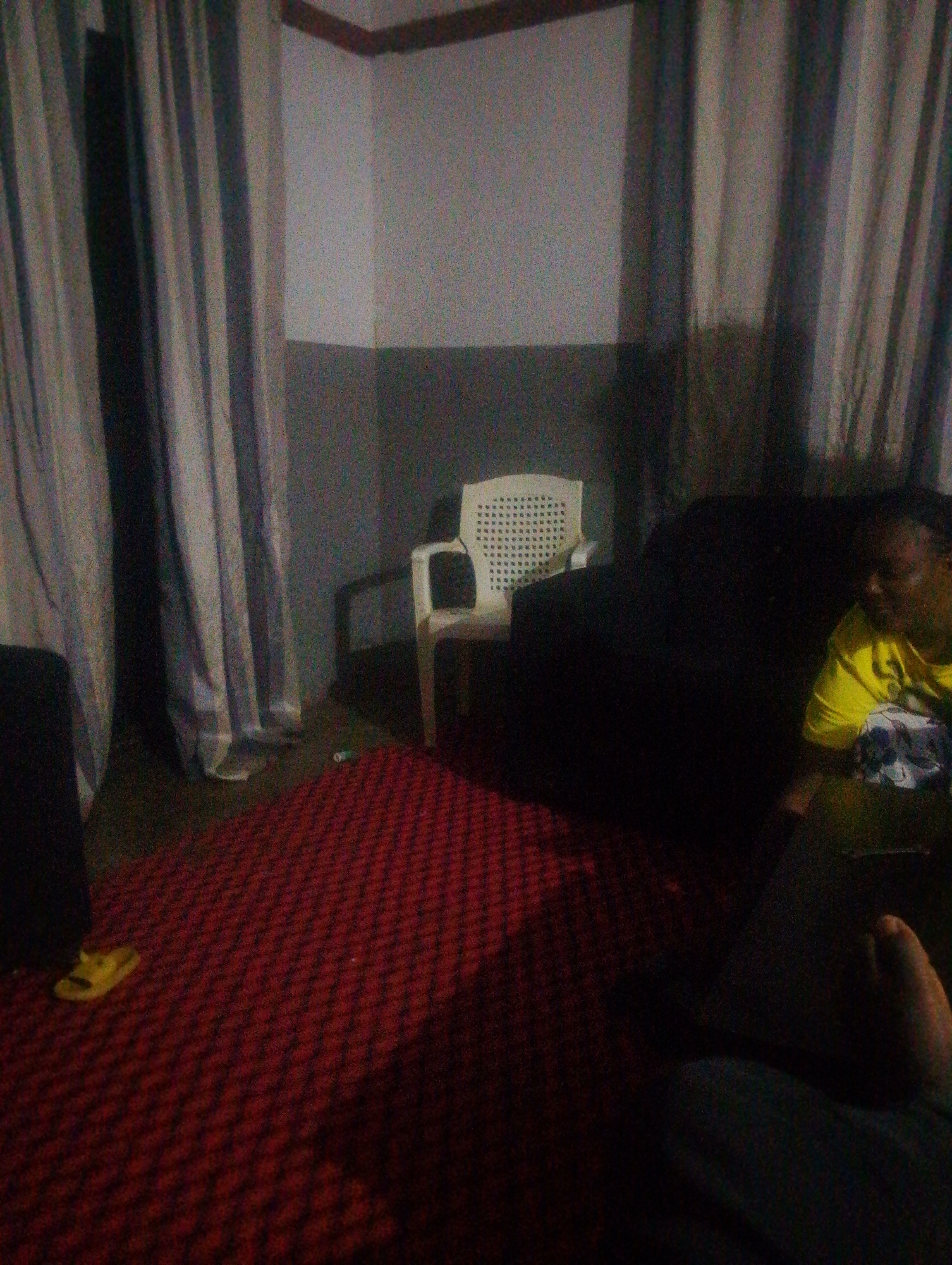 The image depicts an indoor scene, likely a living room or lounge area, with a prominent red patterned carpet covering the floor. To the left, a person is partially visible, wearing yellow footwear. In the background, a white plastic chair is positioned behind a dark sofa. Further to the right, a person in a yellow t-shirt is seated on the sofa. The room is furnished with floor-to-ceiling curtains that appear to be grey and white. The overall lighting is dim, contributing to a grainy texture in the image. The environment suggests a residential setting. The city of Jos, Nigeria, is referenced in the context of this image's potential location, though the image itself does not contain any explicit indicators of its geographical origin within Jos.