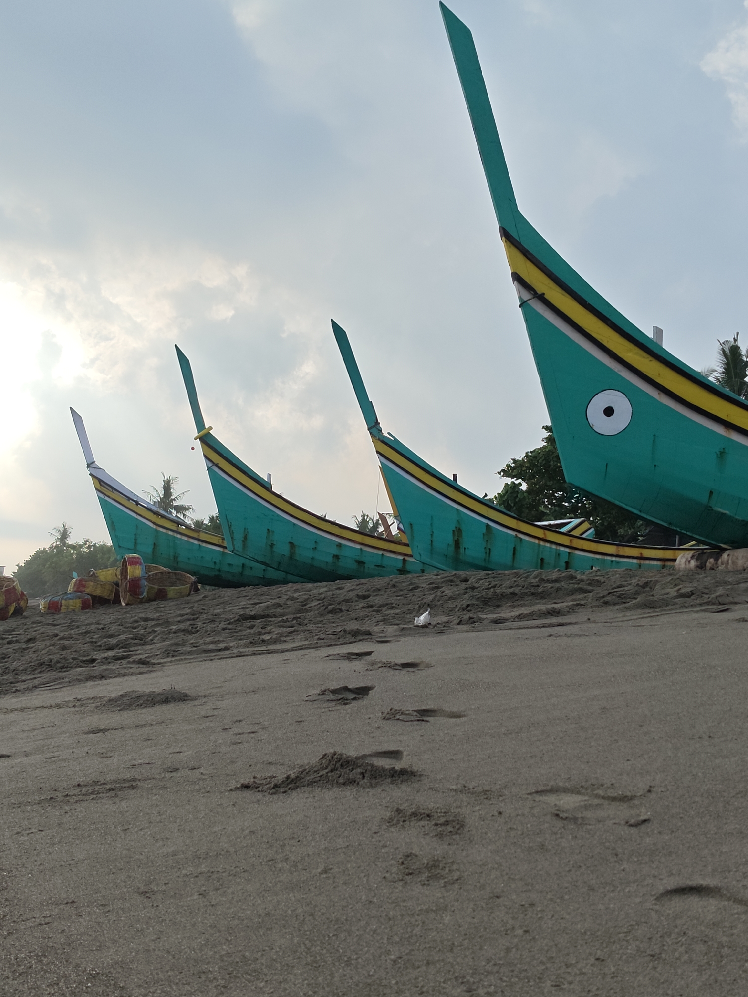 This is a low-angle, eye-level shot of several brightly colored turquoise fishing boats pulled onto a sandy beach. The boats are lined up in a row, facing away from the viewer and towards the sea. They have a distinctive, upward-curving bow, painted with yellow and white stripes. One boat in the foreground has a large, white circular "eye" painted on its side. The sand in the foreground shows numerous footprints and small mounds, suggesting recent activity. In the background, lush green trees and palm trees are visible, along with a hazy sky with scattered clouds. The overall impression is one of a tranquil coastal scene, possibly in a tropical location, in Curehtunong, Indonesia.