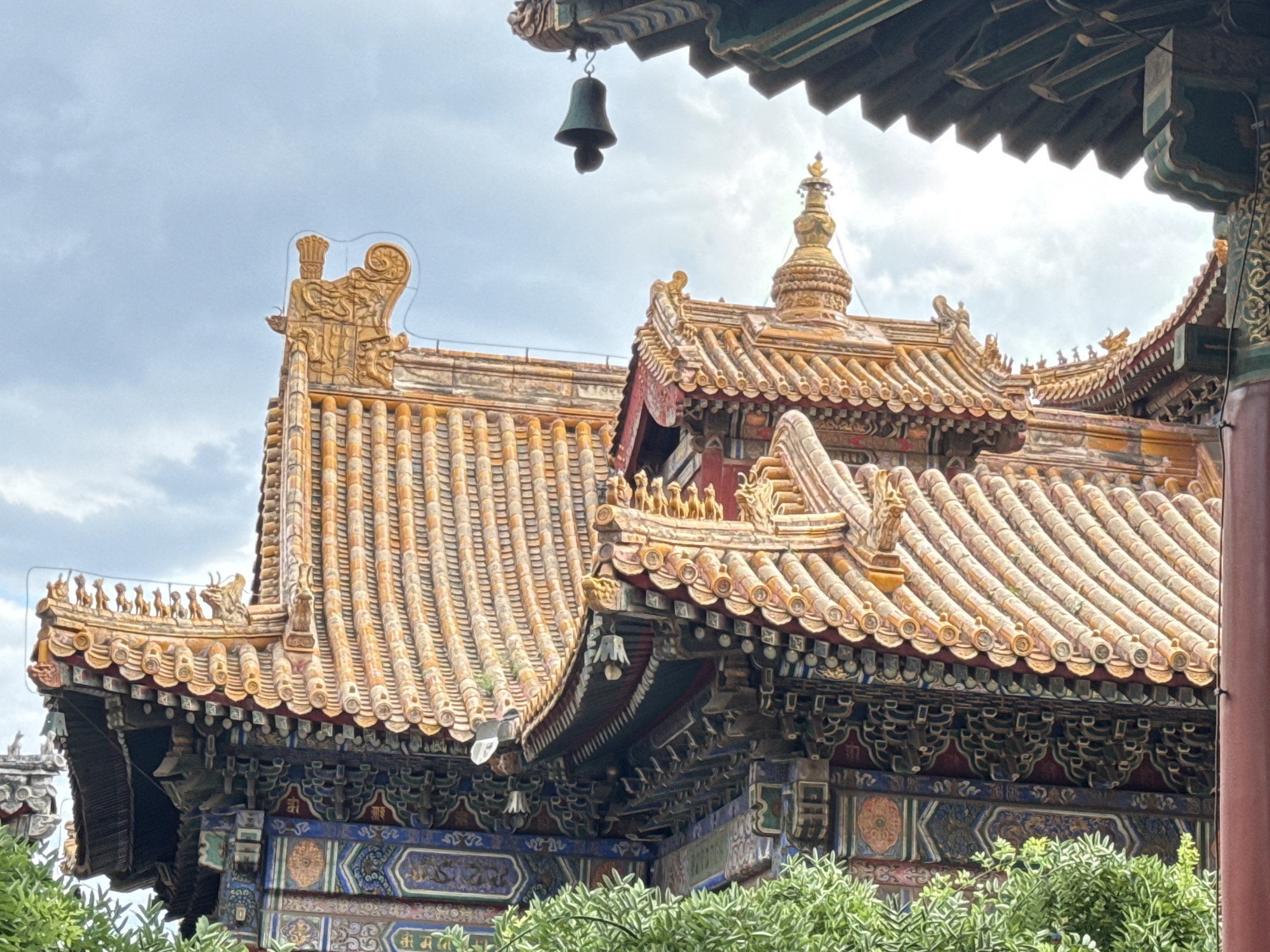 The image shows the ornate, tiled roofs of ancient Chinese architecture, likely in Dongcheng District, China, given the stylistic resemblance to buildings in that area, such as those in the Forbidden City. The roofs are covered in yellowish-orange glazed tiles, with decorative ridges and eaves. Intricate carvings and painted details are visible on the supporting structures beneath the eaves. A dark green bell hangs from the underside of one of the roof structures. In the foreground, lush green foliage obscures the lower parts of the buildings. The sky above is overcast with soft, diffused light. The architectural style suggests a temple or imperial palace.