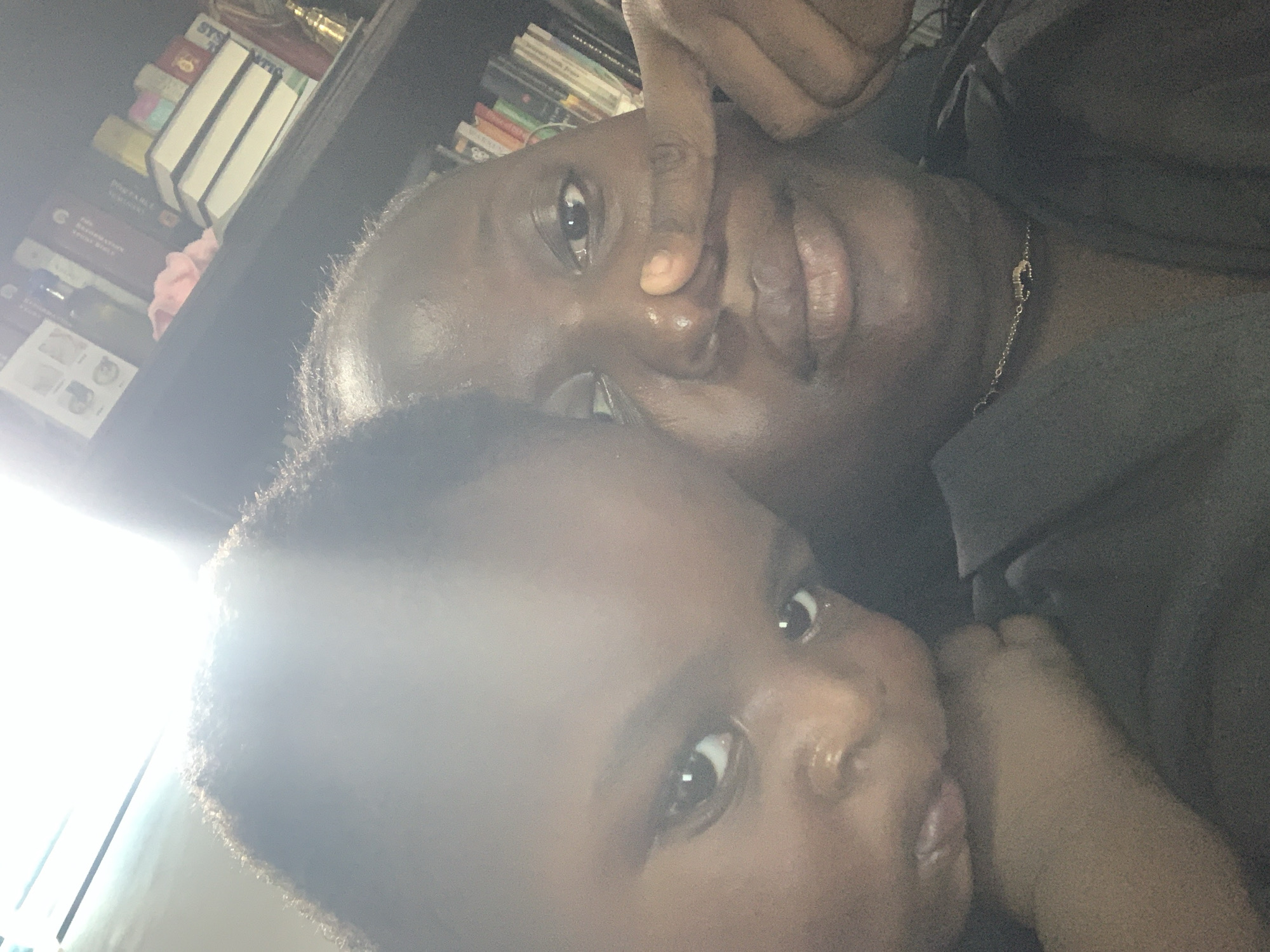 An overhead selfie shows a Black adult and a Black infant. The adult's face is angled slightly upwards and to the right, with a finger touching their nose. The infant's head is tilted to the left and is resting against the adult's face. Both individuals are looking into the camera. Behind them, a bookshelf is visible, filled with books and other decorative items. The lighting is bright, coming from an unseen source above and to the left, creating lens flare and washing out parts of the image.