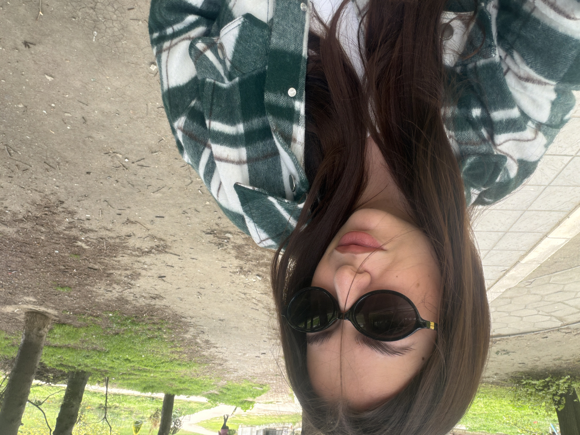 The image is a selfie of a woman with brown hair, wearing black sunglasses and a green and white plaid jacket. She appears to be outdoors in a park or green area. The background shows trees, grass, and a path. The location is likely in the city of Varna, Bulgaria.