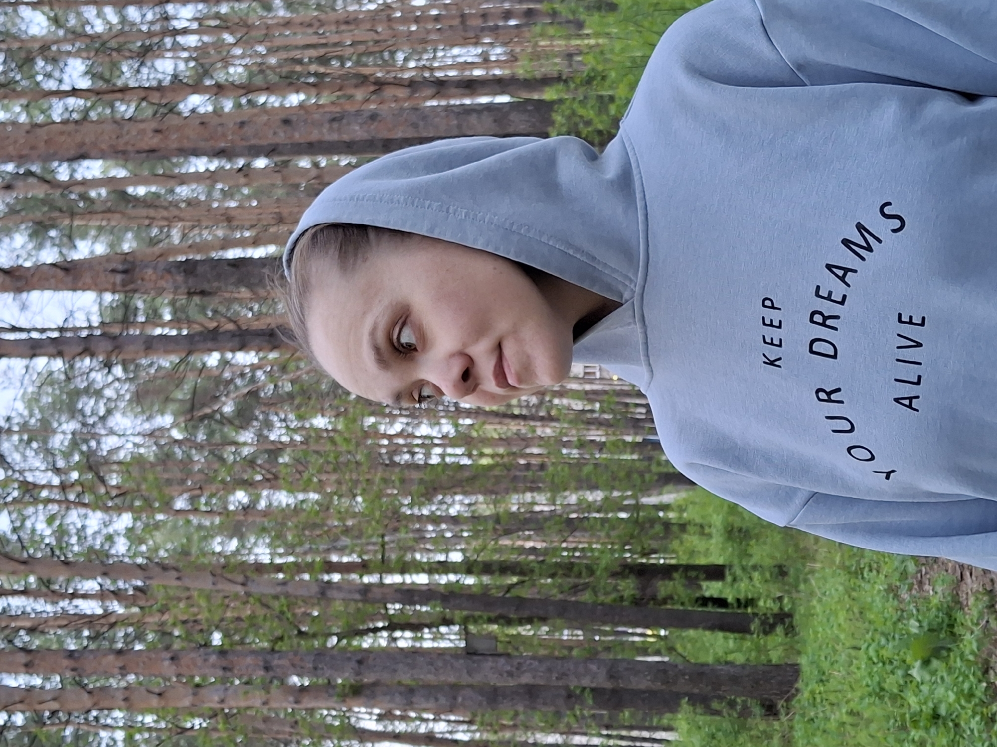 A woman in a light blue hoodie stands in a wooded area. The hoodie has the words "KEEP YOUR DREAMS ALIVE" printed on it in black lettering. The woman's face is visible, and she appears to be looking downwards and slightly to the left. The wooded area consists primarily of tall, slender trees with light-colored bark and green foliage. The background suggests a forest setting within Chaikovsky, Russia.