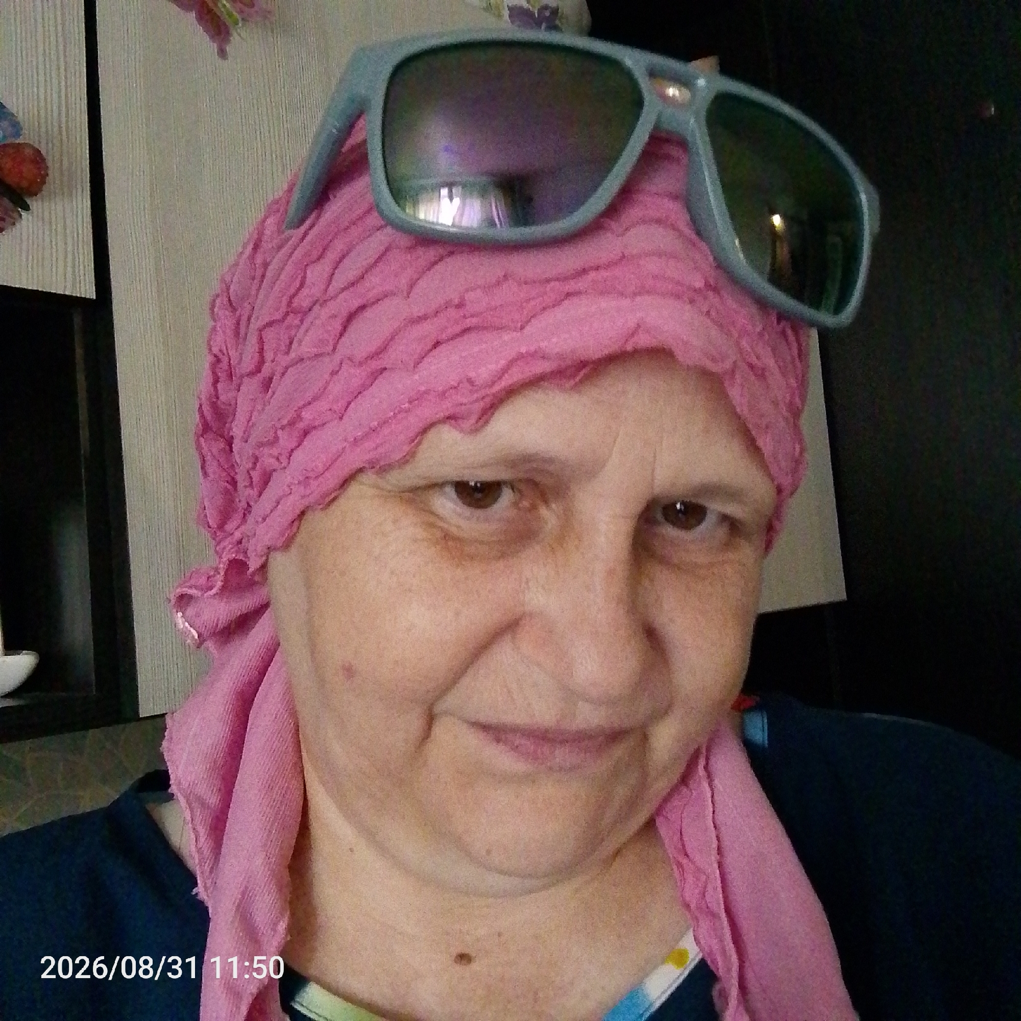 A medium shot selfie shows an adult woman with fair skin, brown eyes, and freckles. She is wearing a pink headscarf that covers her hair and extends around her neck. A pair of light blue sunglasses rests on top of the headscarf. The woman is looking directly at the camera with a slight smile. She is wearing a dark blue top underneath the headscarf. The background is blurred but appears to be indoors, with a wall on the left and a dark surface on the right. A timestamp "2026/08/31 11:50" is visible in the lower left corner of the image.