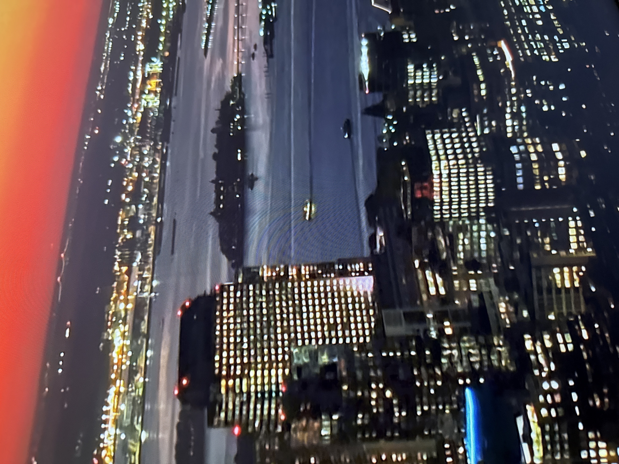 This is a high-angle, night-time aerial photograph of a cityscape at dusk. The sky transitions from a deep red at the top to a dark purple and gray near the horizon, suggesting the sun has recently set or is about to rise. The city lights are prominently displayed, with numerous buildings illuminated by warm yellow and white lights. A body of water, likely a bay or river, is visible in the midground, reflecting the city lights.
Several boats or ships are present on the water. In the foreground, large, illuminated buildings dominate the scene, showcasing their grid-like patterns of windows. One particularly tall building in the center has red lights at its top. The overall impression is of a bustling urban environment at night. The specific location is not identifiable as Burgas, Bulgaria based on visual evidence within the image.