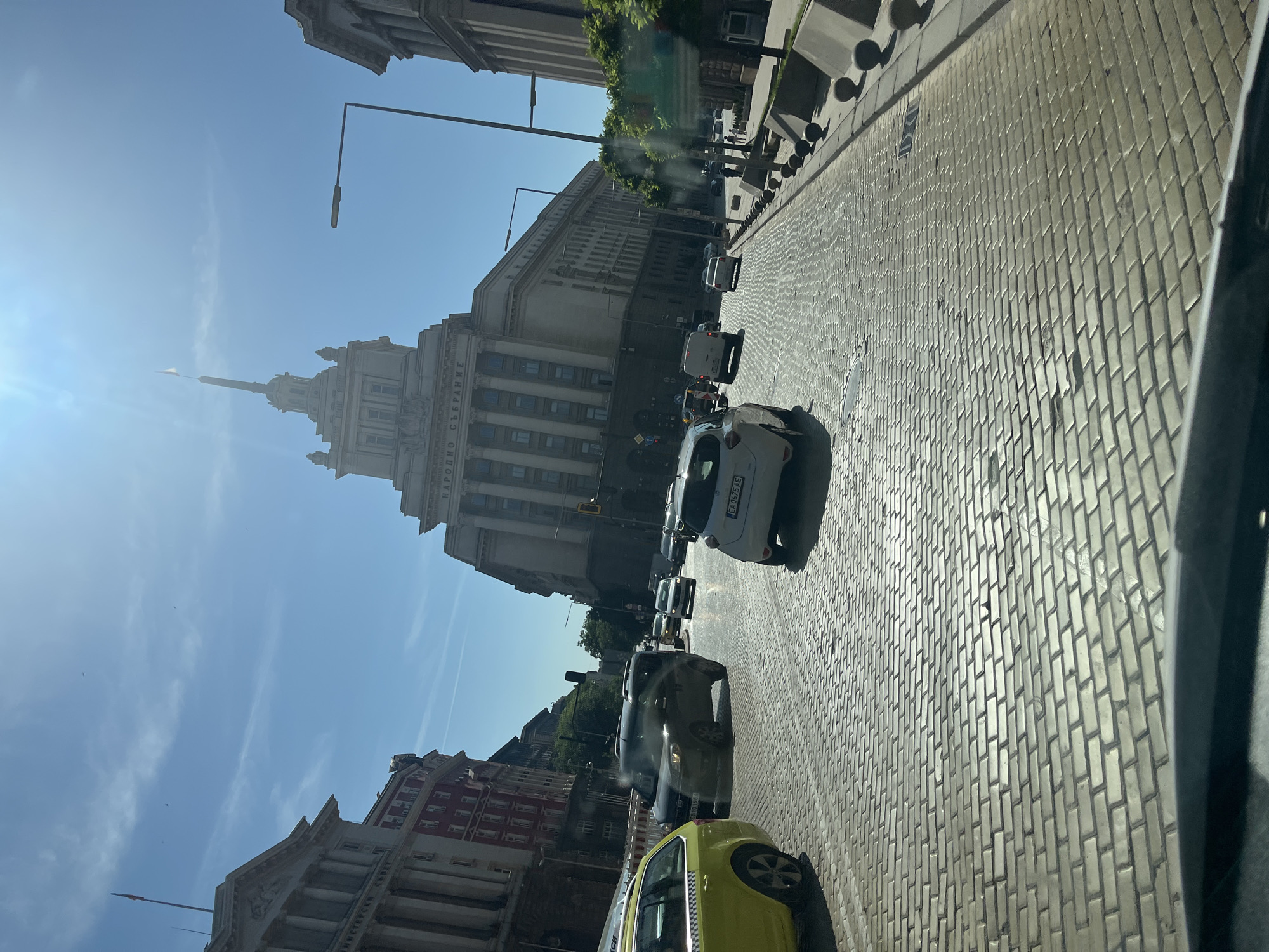 The image depicts a street scene in a city under a clear blue sky with the sun shining brightly. The foreground is dominated by a cobblestone road, reflecting the sunlight. Several cars are visible on the road, including a yellow taxi on the left, a dark-colored minivan in the middle, and a white car in the foreground. In the background, a grand, multi-story building with classical architecture stands prominently, featuring columns and a tall spire topped with a flag. The building has the inscription "НАРОДНО СЪБРАНИЕ" visible on its facade. Adjacent to it, another large building with similar architectural style is partially visible. Streetlights and traffic signals are also present. The scene suggests a bustling urban environment, possibly a capital city, given the monumental architecture and the inscription on the building, which translates to "National Assembly."