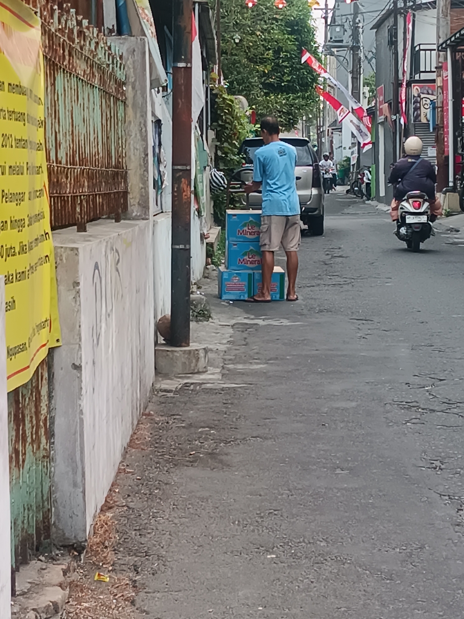 The image captures a street scene in the city of Yogyakarta, Indonesia. In the foreground, a person wearing a blue t-shirt and khaki shorts is standing next to several cases of bottled water, identified by the "Le Mineral" branding. The person is wearing flip-flops. The cases of water are stacked on the asphalt street. Behind the person, a silver car is parked. Further down the street, two individuals are riding on a scooter, with the rider in the front wearing a white helmet and the passenger behind wearing a dark jacket and helmet. Red and white Indonesian flags are displayed on poles along the street, suggesting a national celebration or event. The buildings lining the street appear to be a mix of residential and commercial structures, with some showing signs of age. A rusty metal fence is visible on the left side of the image, alongside a yellow banner with Indonesian text. The overall environment suggests a moderately busy urban area, possibly during the daytime.