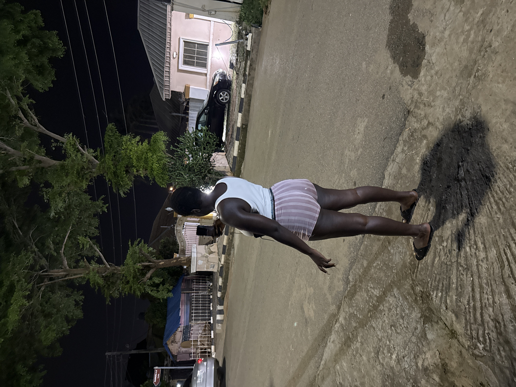 A person stands on a street in Abuja, Nigeria, at night, holding a smartphone. The person is wearing a white tank top and striped shorts. In the background, there are houses, trees, and cars. The street is paved and appears wet in some areas, reflecting the artificial light. The overall impression is that of a common urban scene at night in the city.