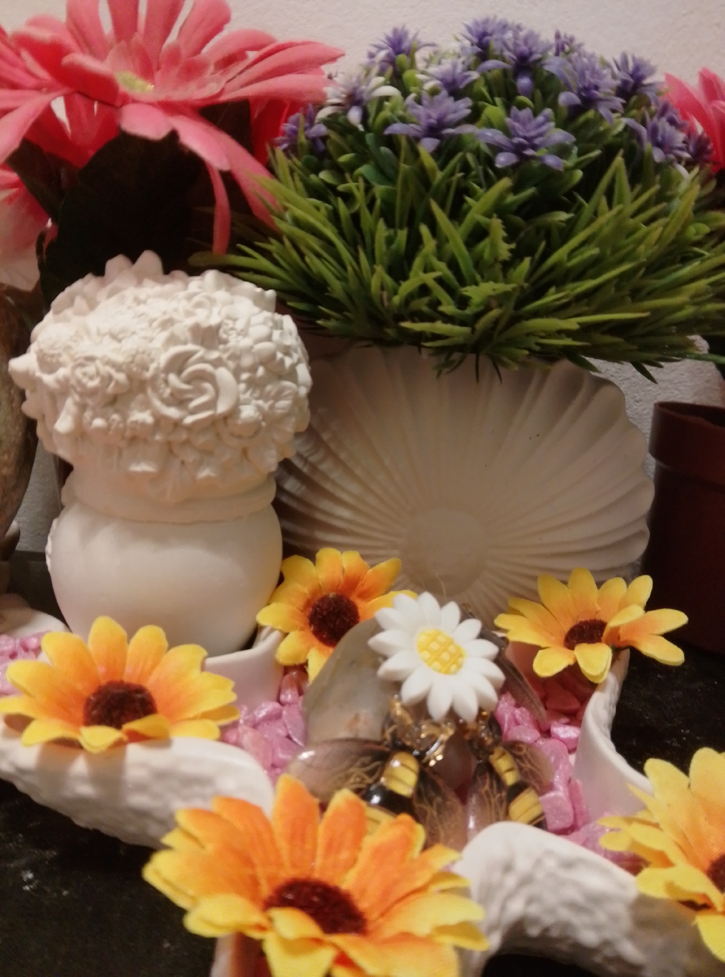 The image depicts a close-up view of a decorative arrangement of artificial flowers and figurines. In the foreground, there are several bright yellow and orange artificial sunflowers with dark centers. Some are arranged in white decorative shells. Towards the center of the arrangement, a small white figurine of a daisy-like flower with a yellow center is visible, along with decorative bee figurines. Behind these, a white ceramic vase shaped like a shell holds a cluster of artificial greenery with small purple flowers. To the left of this arrangement, a portion of a large pink artificial flower is visible. The background shows a light-colored wall and a brown terracotta pot. The overall scene appears to be an indoor decorative display. There is no indication of the city of Wedel, Germany, or any specific location within it.