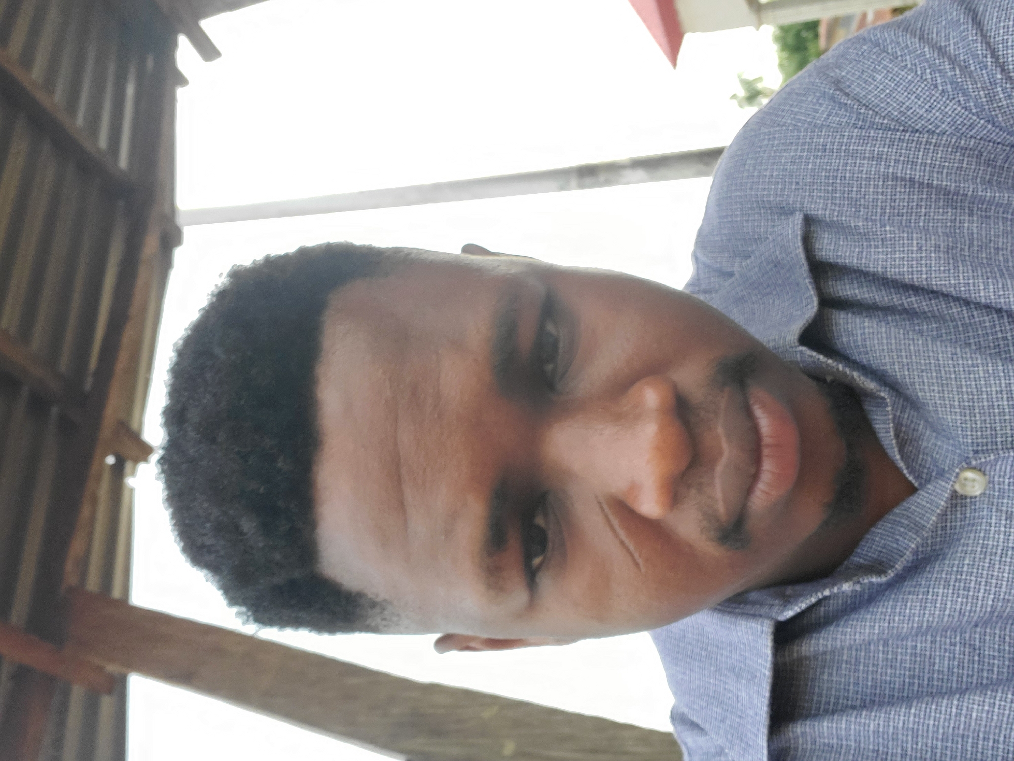 A close-up portrait of a young Black man is depicted, rotated 90 degrees counter-clockwise. The man has short, dark, curly hair and a short beard or stubble. He wears a blue collared shirt with a subtle checked or plaid pattern and a visible white button near his chest. He looks directly at the viewer with a friendly, engaging smile.
The background is highly overexposed and bright, indicative of strong daylight. On the left side, dark, rustic architectural elements, possibly wooden beams or corrugated metal sheets forming a ceiling or wall, are visible, suggesting a sheltered outdoor setting or a simple building structure. A light-colored horizontal bar crosses the mid-ground. In the upper right corner, hints of a red architectural feature, possibly a roof edge, and some green foliage are discernible. The lighting is bright, illuminating the subject's face well against the bright backdrop. The image conveys a casual and approachable mood, likely captured during the daytime in Mararaba, Nigeria. There is no visible text in the image.