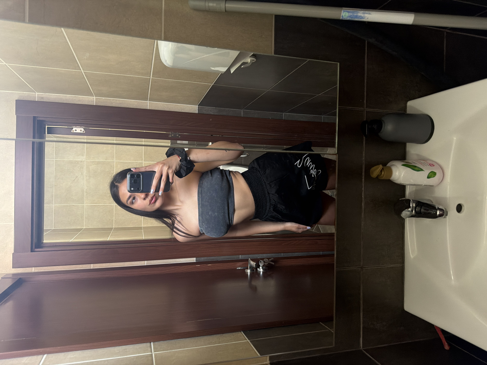 A young woman is captured in a mirror selfie inside a bathroom, located in Gabrovo, Bulgaria. The image is rotated 90 degrees counter-clockwise.
The woman is the central subject, holding a black iPhone in her right hand to take the photo. She has long, dark hair and is wearing a gray, ribbed bandeau top and black athletic shorts with white stylized text, partially visible as "Love." A black scrunchie is on her left wrist, and her fingernails have light-colored polish.
The bathroom features light brown rectangular tiles on the upper walls and dark gray square tiles on the lower walls and floor. A dark brown wooden door frame and door are prominently reflected in the mirror. To the right, a white sink is visible, holding a dark gray pump dispenser and a white Dove brand lotion bottle with green accents, displaying the word "DOVE" and partially legible "GENTLE." In the upper reflected area, a white toilet and a gray pipe with an illegible blue and white sticker can be seen. A set of keys is also reflected on a surface near the bottom of the mirror. The lighting suggests an indoor setting, likely artificial. The woman's expression is neutral as she focuses on her phone.