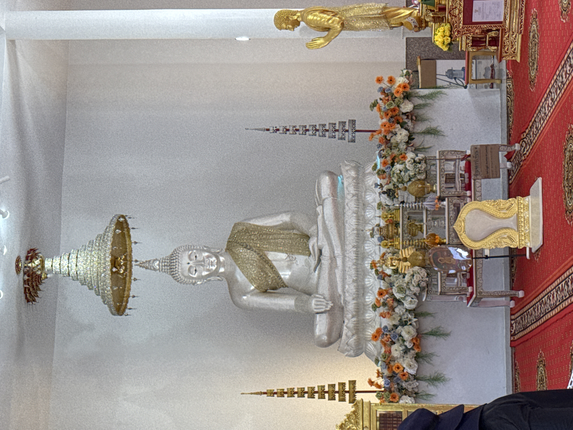 The image shows an interior scene within what appears to be a Buddhist temple in Thep Krasatti, Thailand. Dominating the center is a large, seated, silver Buddha statue on a lotus pedestal, adorned with a golden sash. Above the Buddha statue, a tiered, ornate umbrella-like structure hangs from the ceiling. To the right of the Buddha statue stands a smaller, golden statue of a figure with hands pressed together in a prayerful gesture. In front of the Buddha, a tiered altar is decorated with an abundance of white, blue, and orange flowers, along with various golden ceremonial objects. To the left of the Buddha statue, a tall, golden tiered structure with intricate carvings is visible. In the foreground, a red carpet with a black and white patterned border covers the floor. A person, wearing a dark blue headdress and black clothing, is partially visible on the far left of the frame. The environment is well-lit with a white wall in the background.